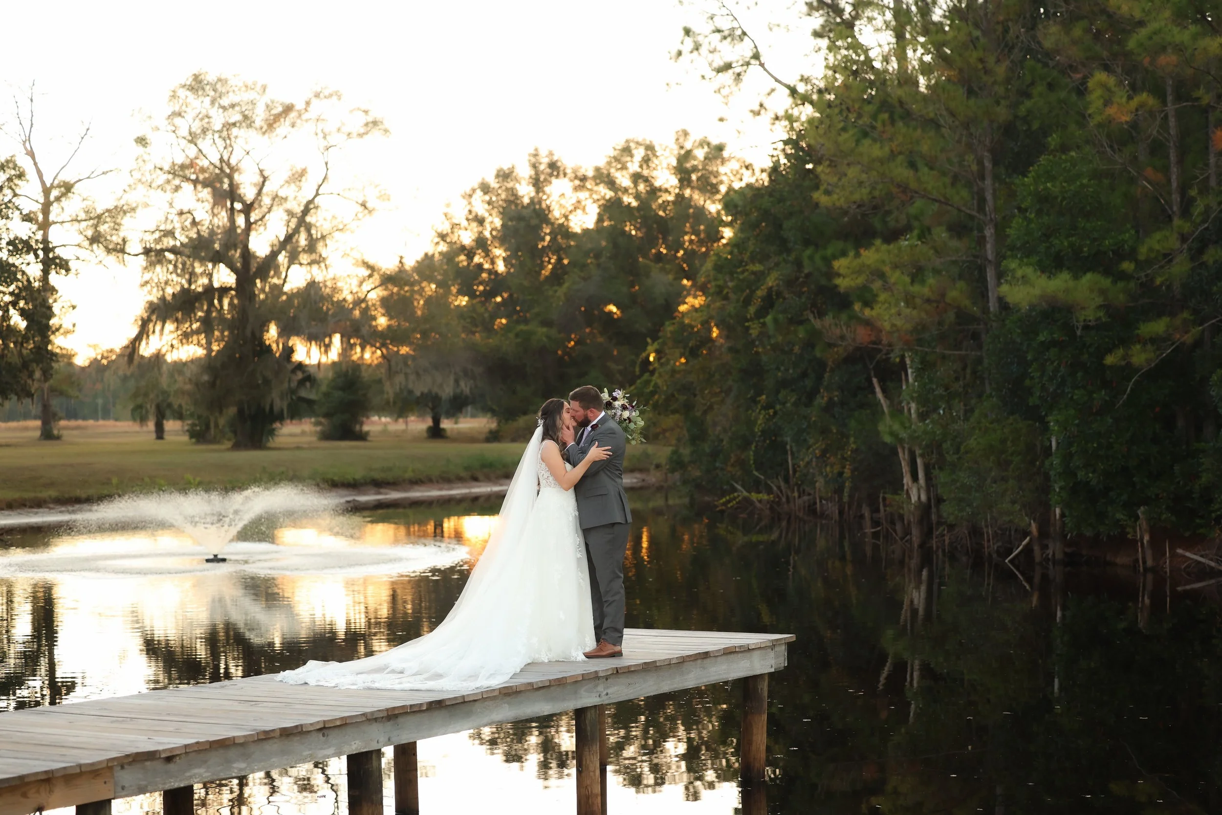 Bride and groom embracing on a dock at sunset, reflected in the water with trees and soft evening light behind them.