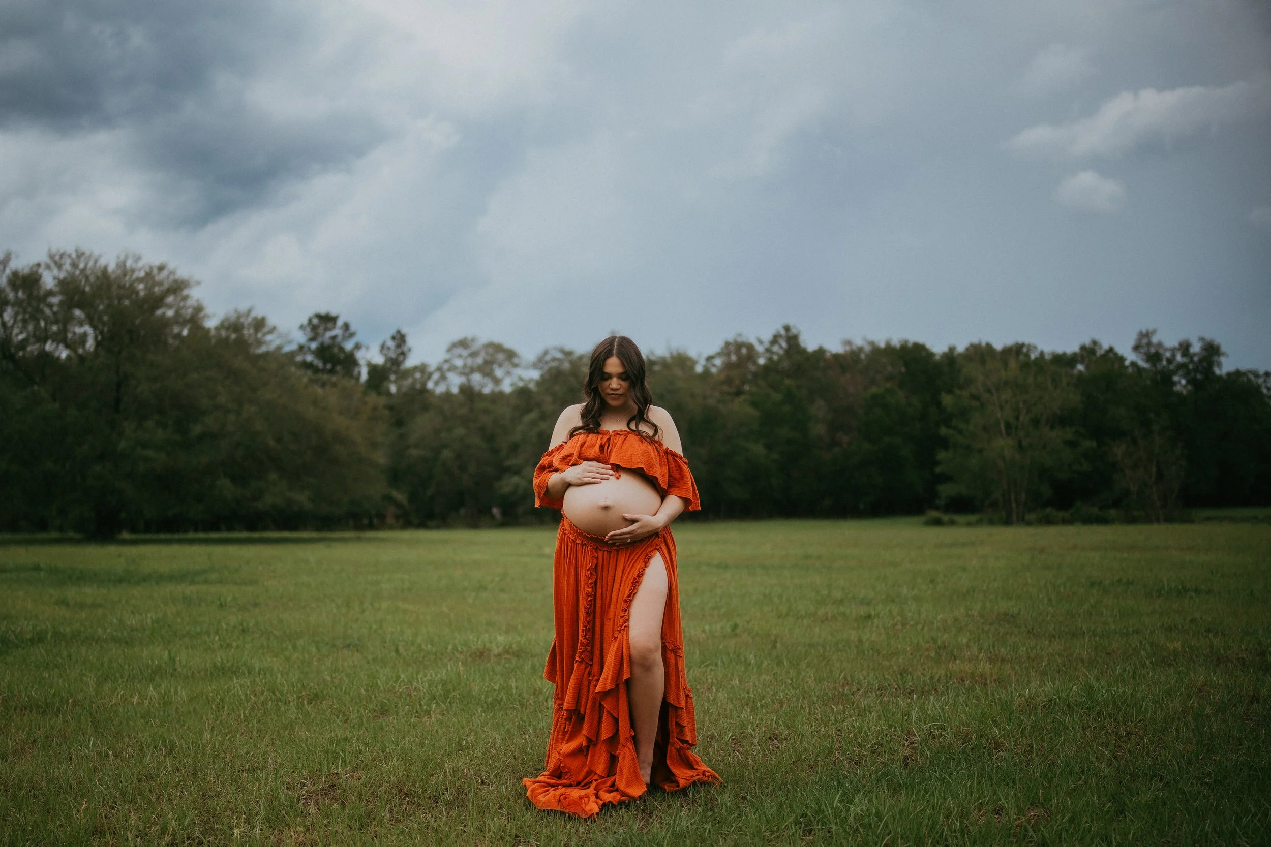 Expectant mother cradles her baby bump in a grassy field beneath dramatic skies during a maternity session in Hilliard, Florida.