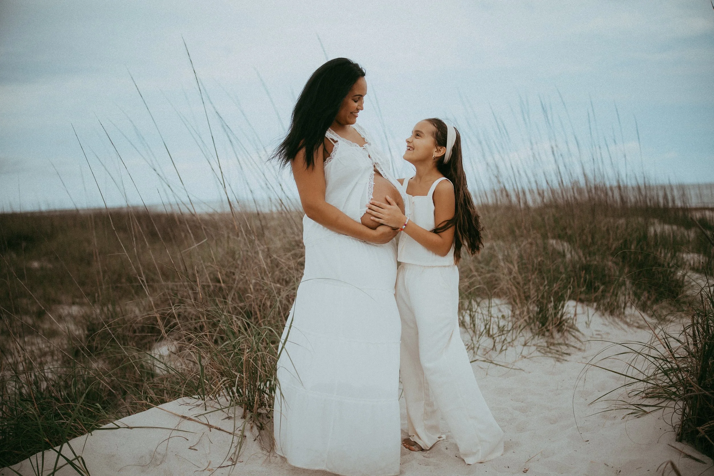 Expectant mother and her daughter stand together among the sea oats at Fort Clinch State Park in Fernandina Beach, Florida, smiling at one another while gently holding the baby bump during a beach maternity photography session.