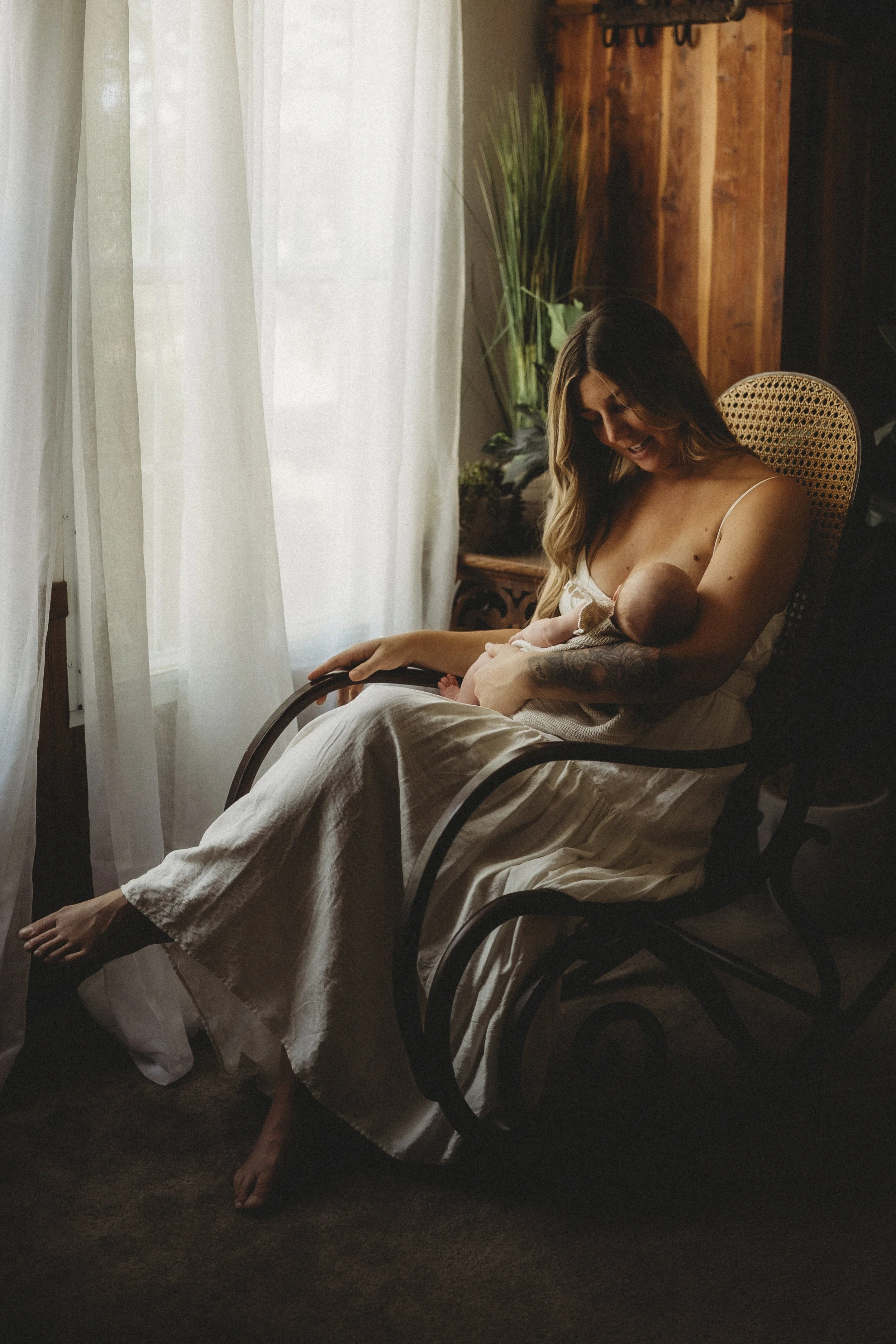 Mother resting in a rocking chair by a sunlit window, holding her newborn close as gentle light fills the quiet room.