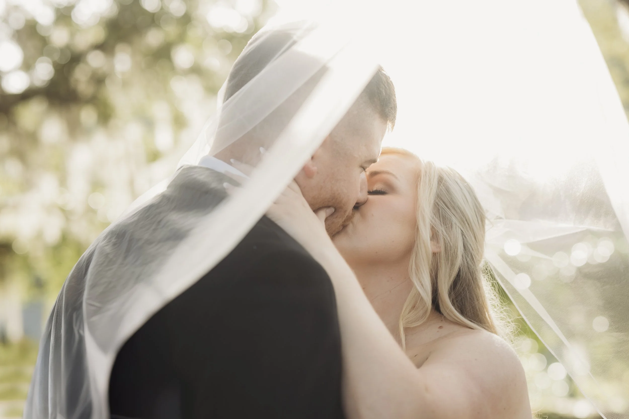 Sunlight filters through her veil as they kiss, a tender moment suspended in warmth and promise.