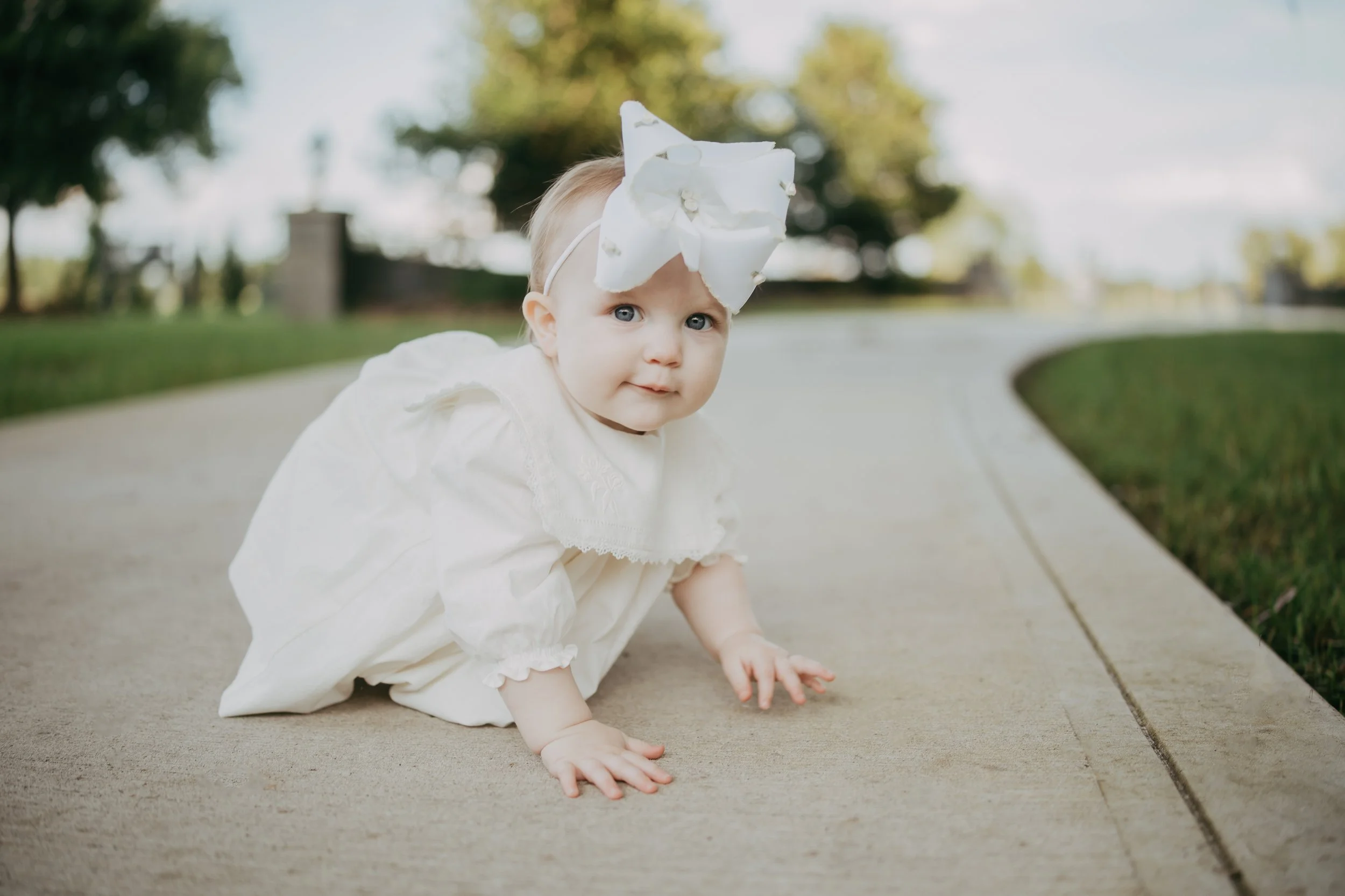 A curious baby girl in a white heirloom dress and oversized bow crawls along a quiet country road, creating a sweet milestone portrait filled with innocence and timeless charm.