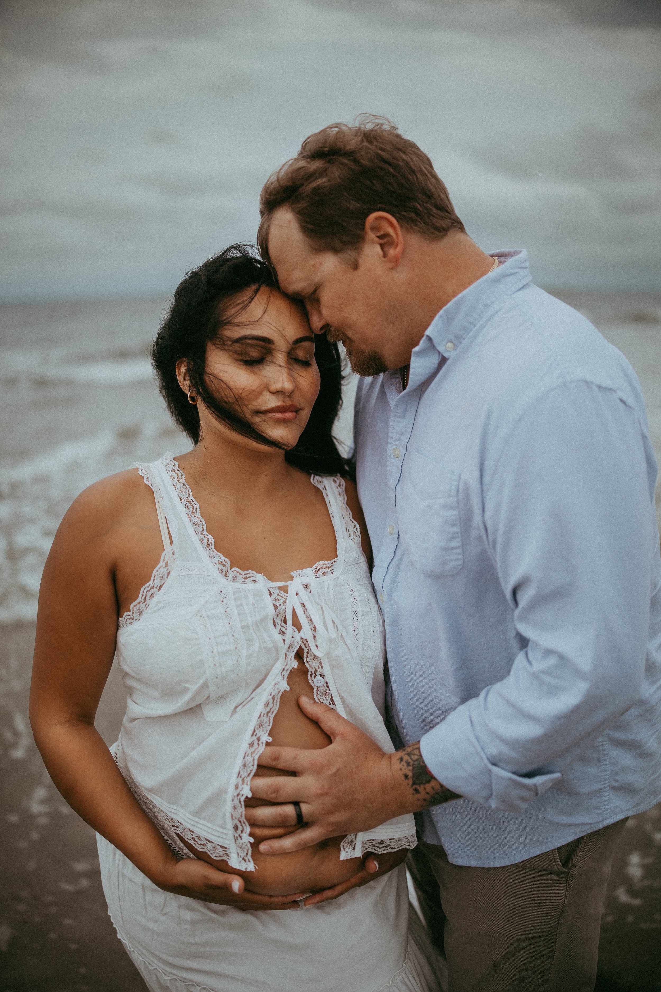 A couple holding a pregnant woman on a beach, with their foreheads touching, embracing her belly and expressing affection.