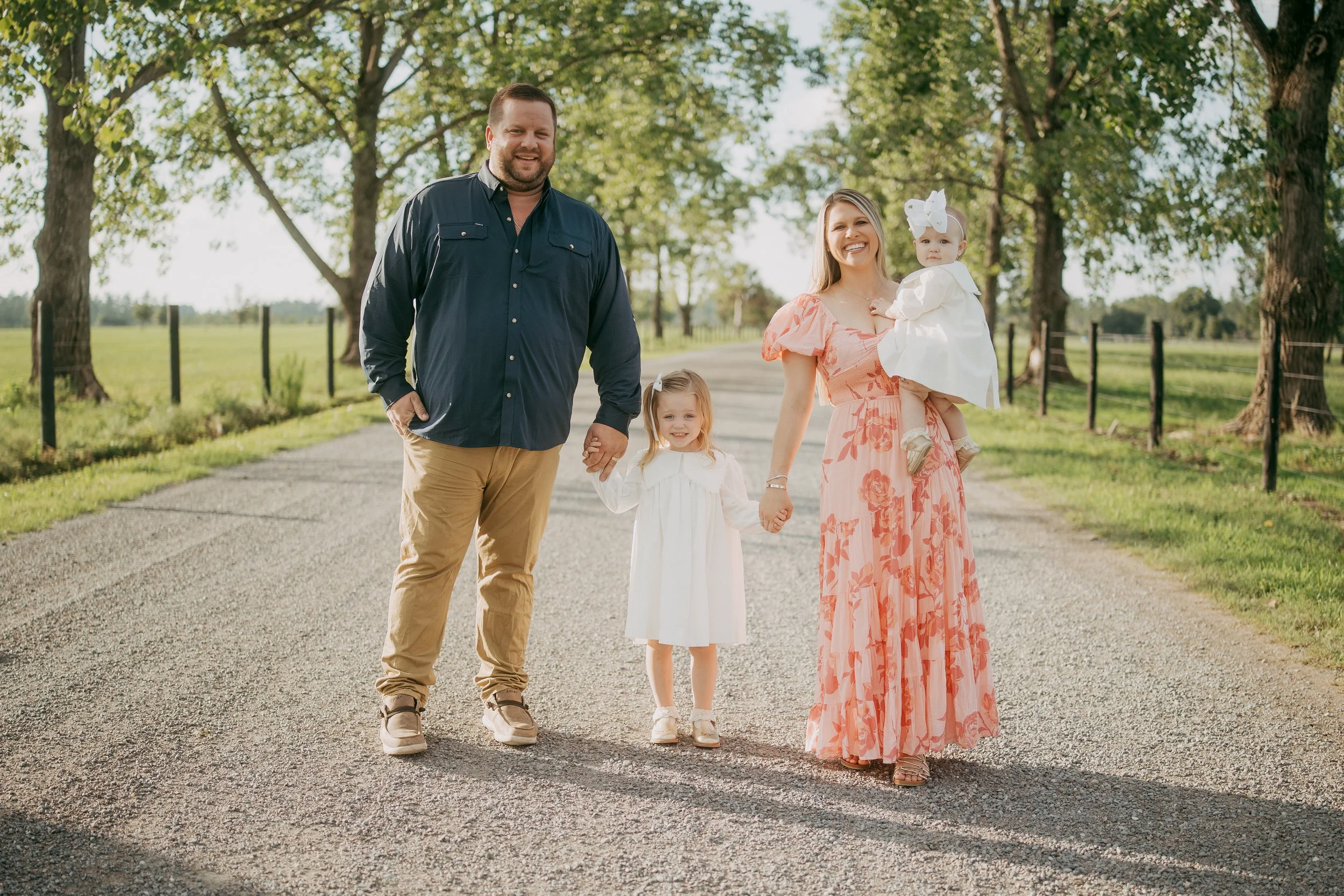 A joyful family of four walks together down a sunlit country road, surrounded by towering trees and soft golden light, creating a timeless outdoor family portrait.
