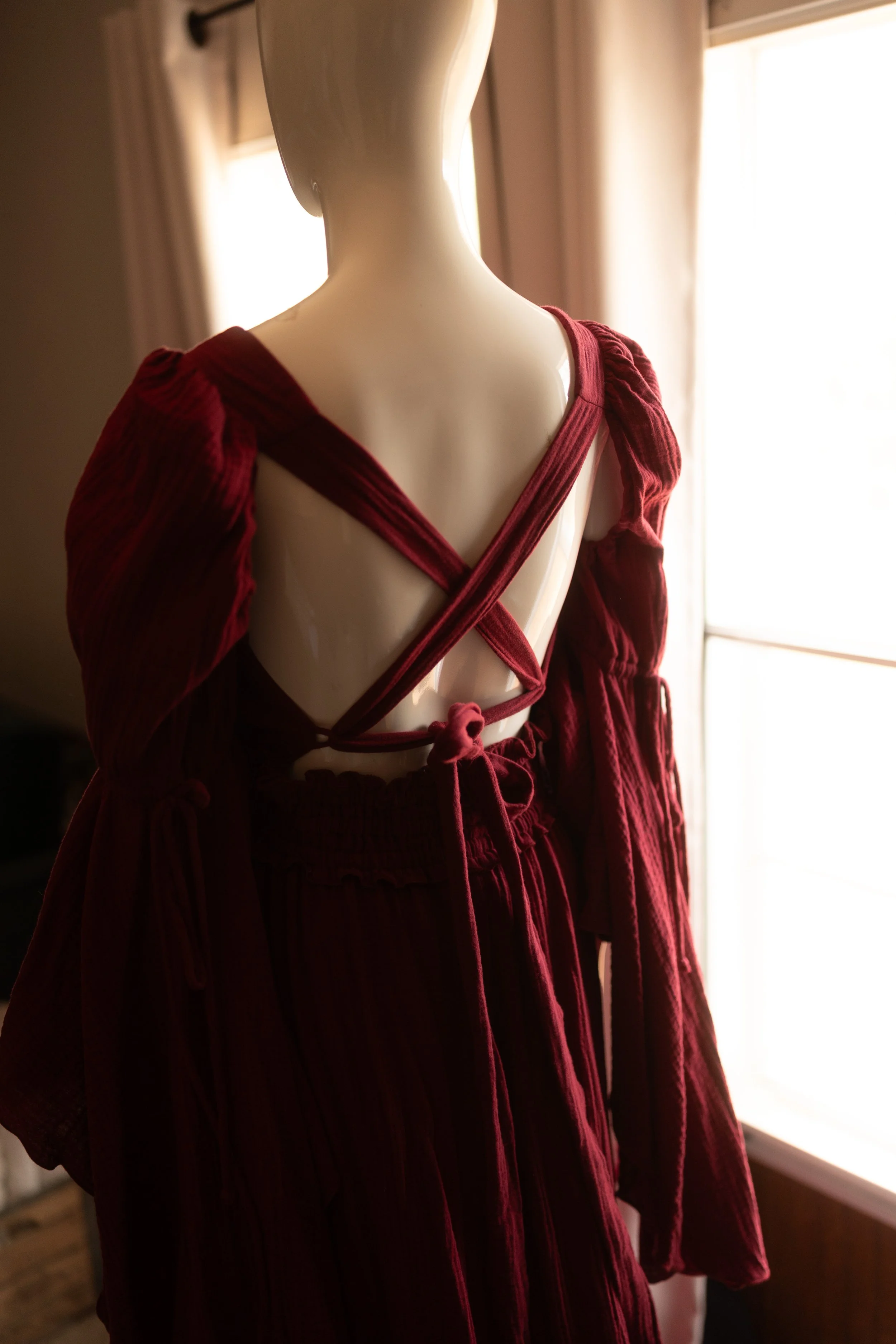 Deep burgundy maxi dress with long billowing sleeves, a V-neck bodice, and a crisscross open back with tie detail, styled on a mannequin in warm natural light.