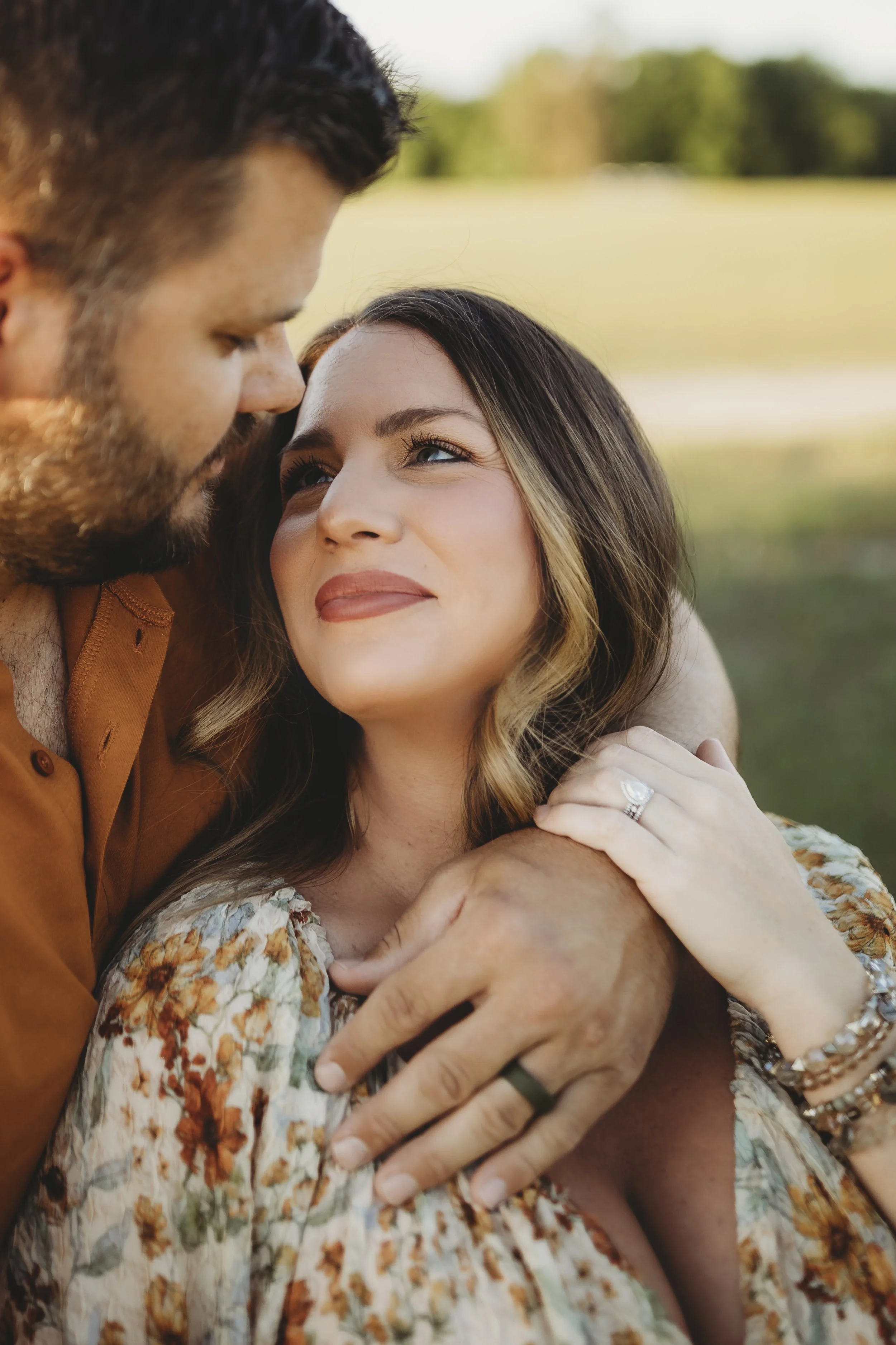Intimate maternity portrait of an expecting couple in a sunlit field, with the father’s arms wrapped around the mother as they gaze at each other.