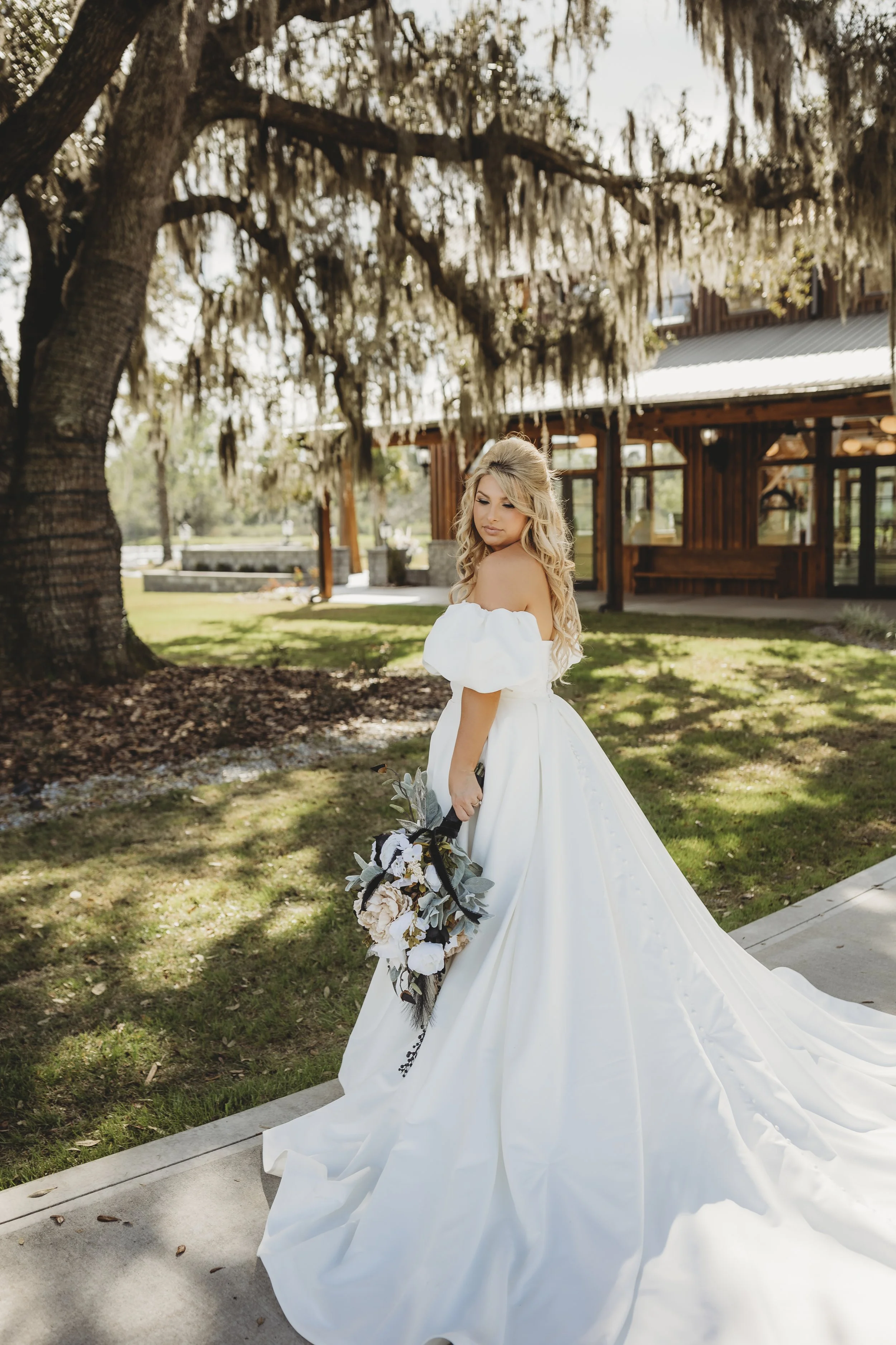 A woman in a white wedding dress holding a bouquet of flowers outdoors under a large tree with hanging moss, in front of a wooden building.