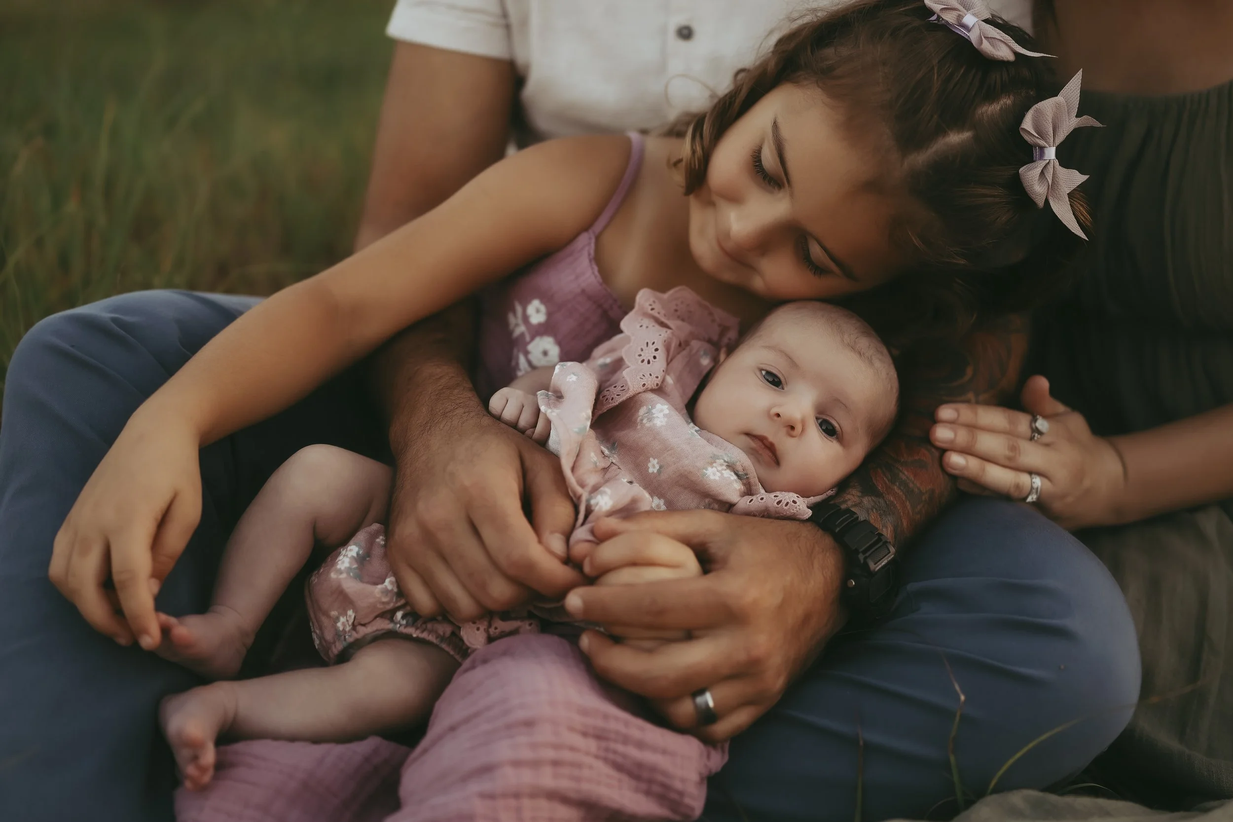 A big sister cuddles her newborn sibling while surrounded by the loving embrace of her family during a sunset session.