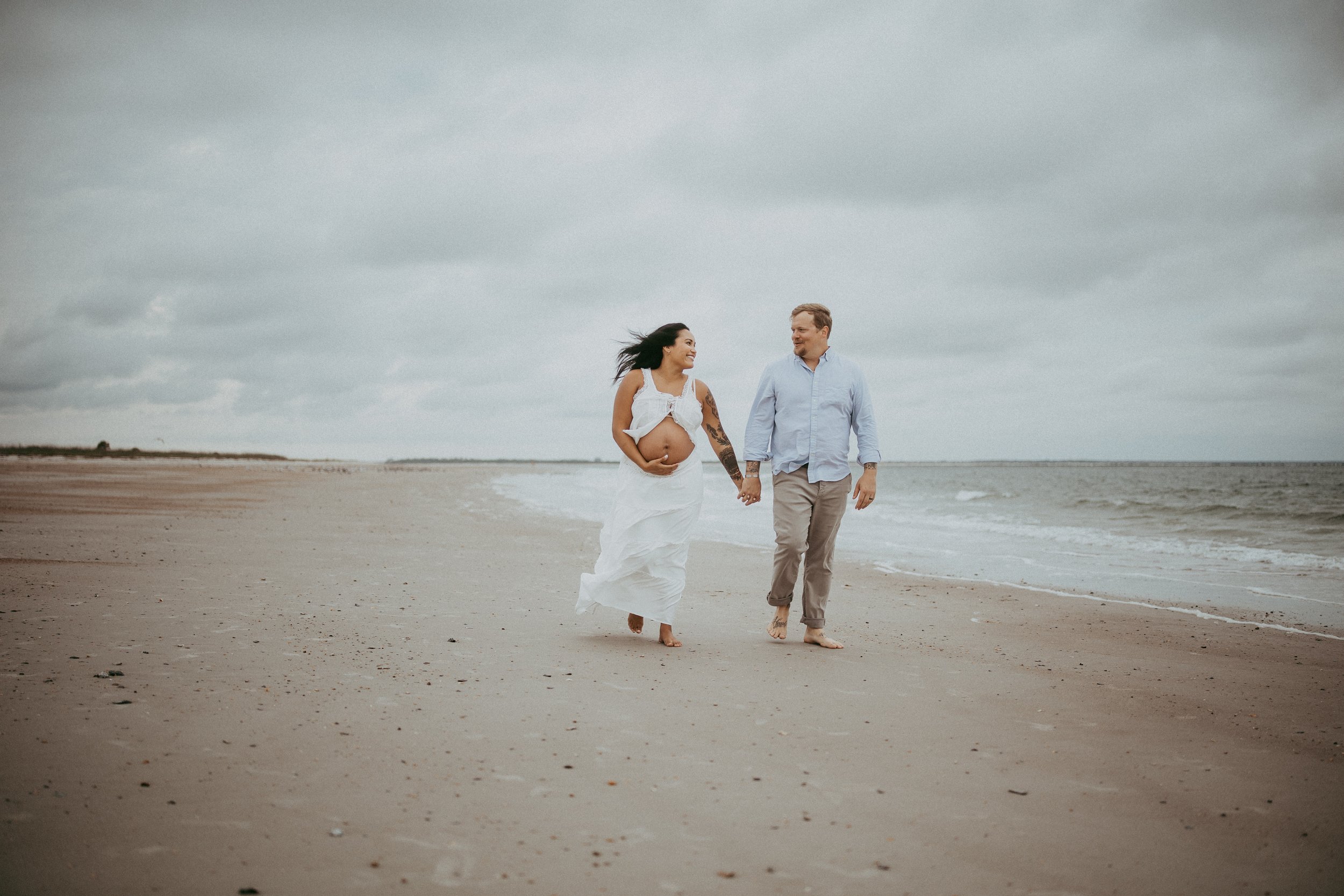Pregnant couple walks hand in hand along the shoreline at Fort Clinch State Park in Fernandina Beach, Florida, sharing smiles beneath a dramatic cloudy sky during a relaxed beach maternity photography session.