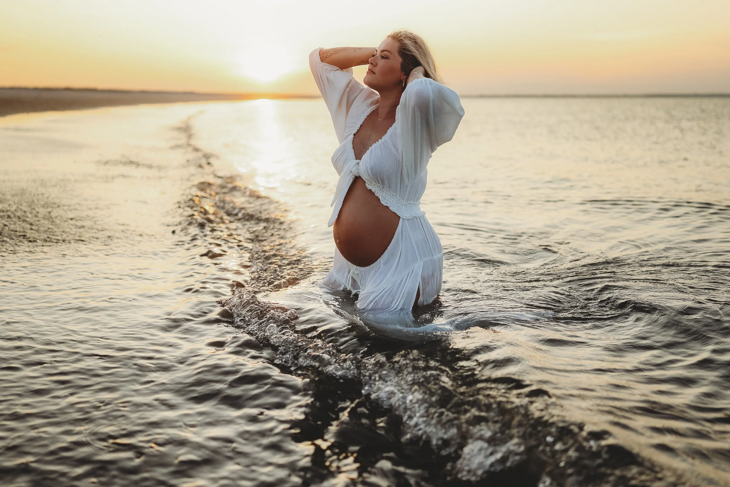 Sunset beach maternity photo of an expectant mother in a white dress, standing in the ocean with her hands in her hair as golden light reflects across the water.