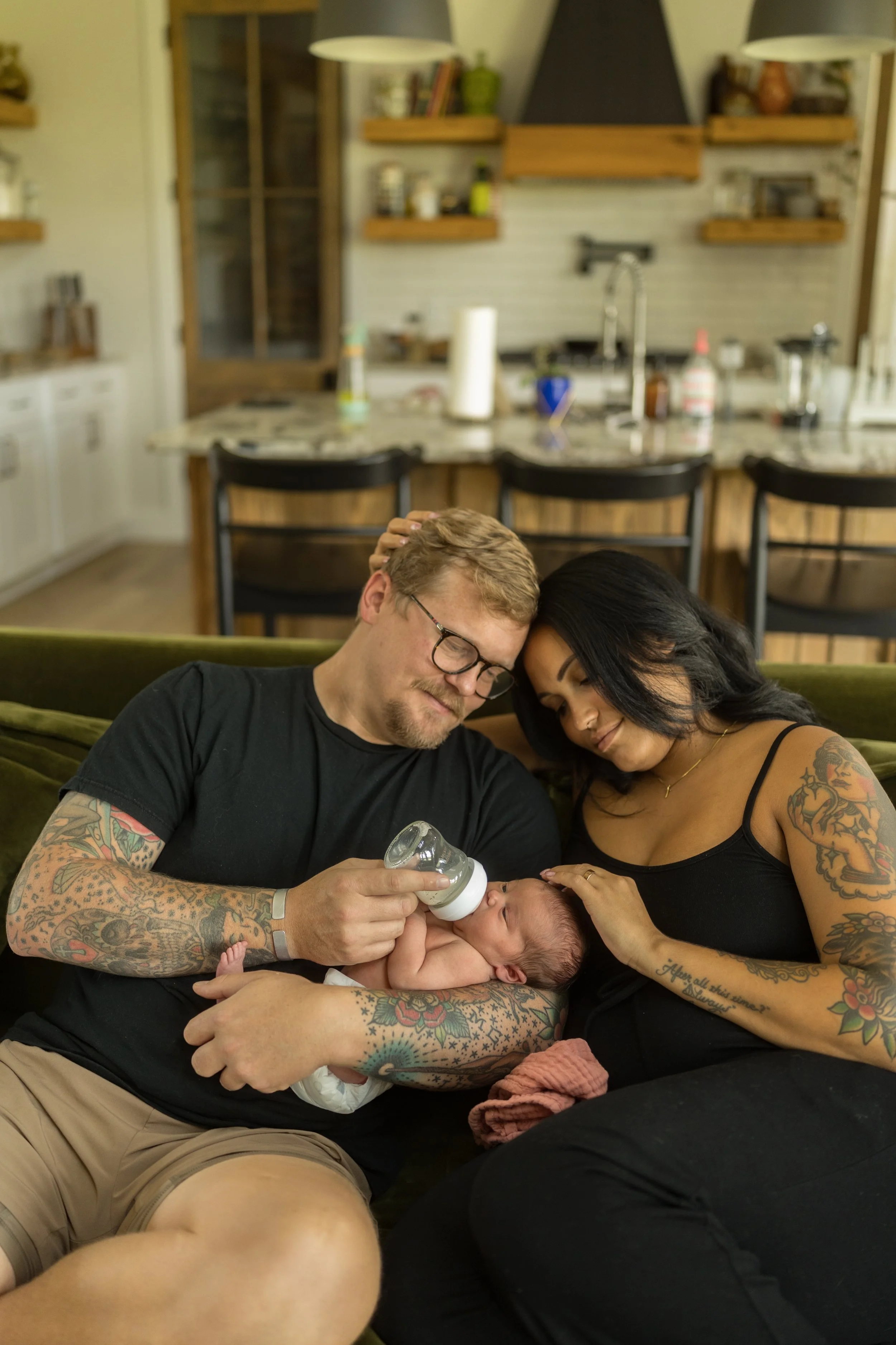Father bottle-feeds his newborn while mother gently strokes the baby's head, sharing a quiet everyday moment together on the living room sofa during an in-home lifestyle newborn photography session.