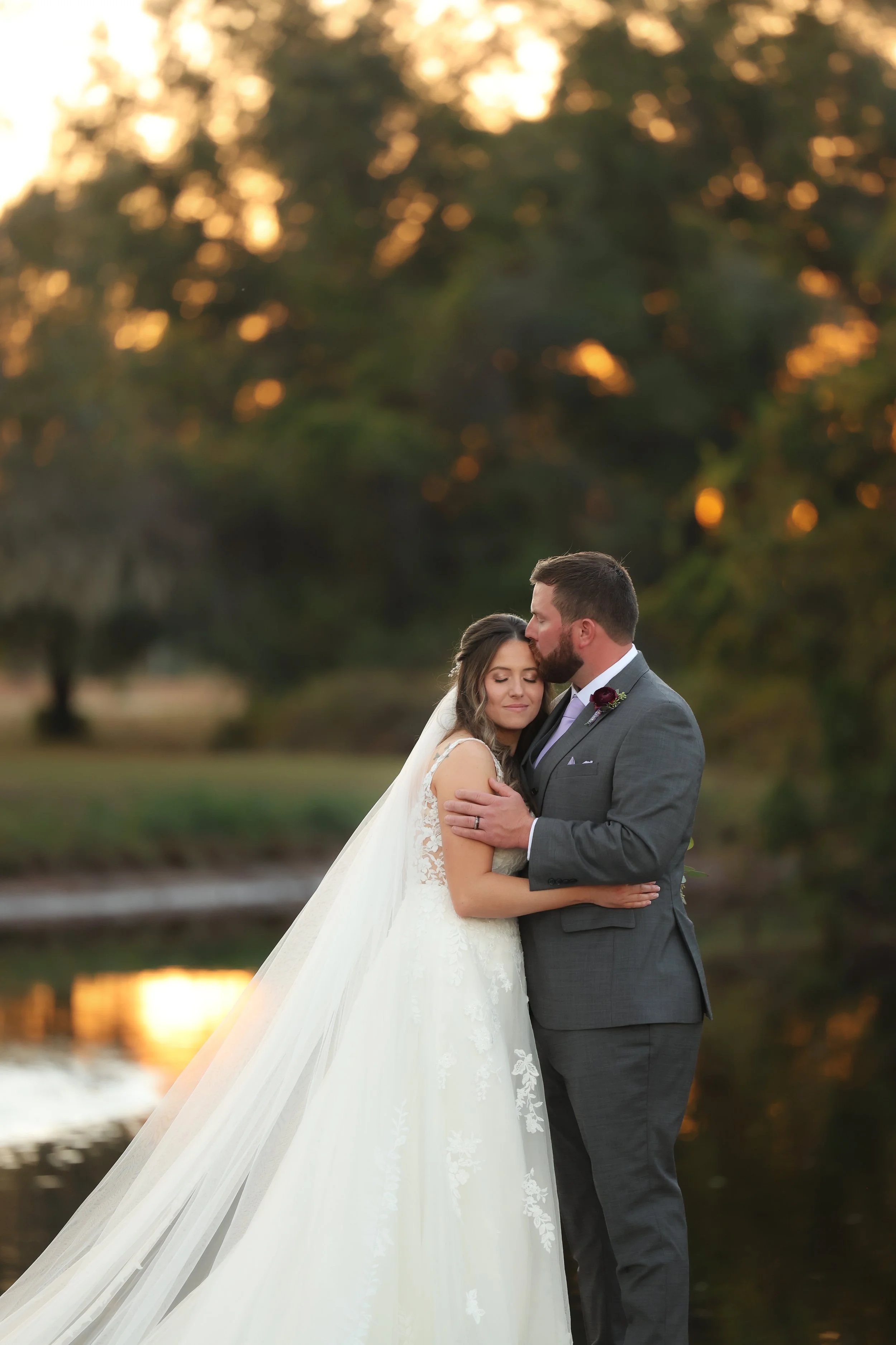 Groom gently kissing the bride’s forehead as they embrace by the water at sunset.