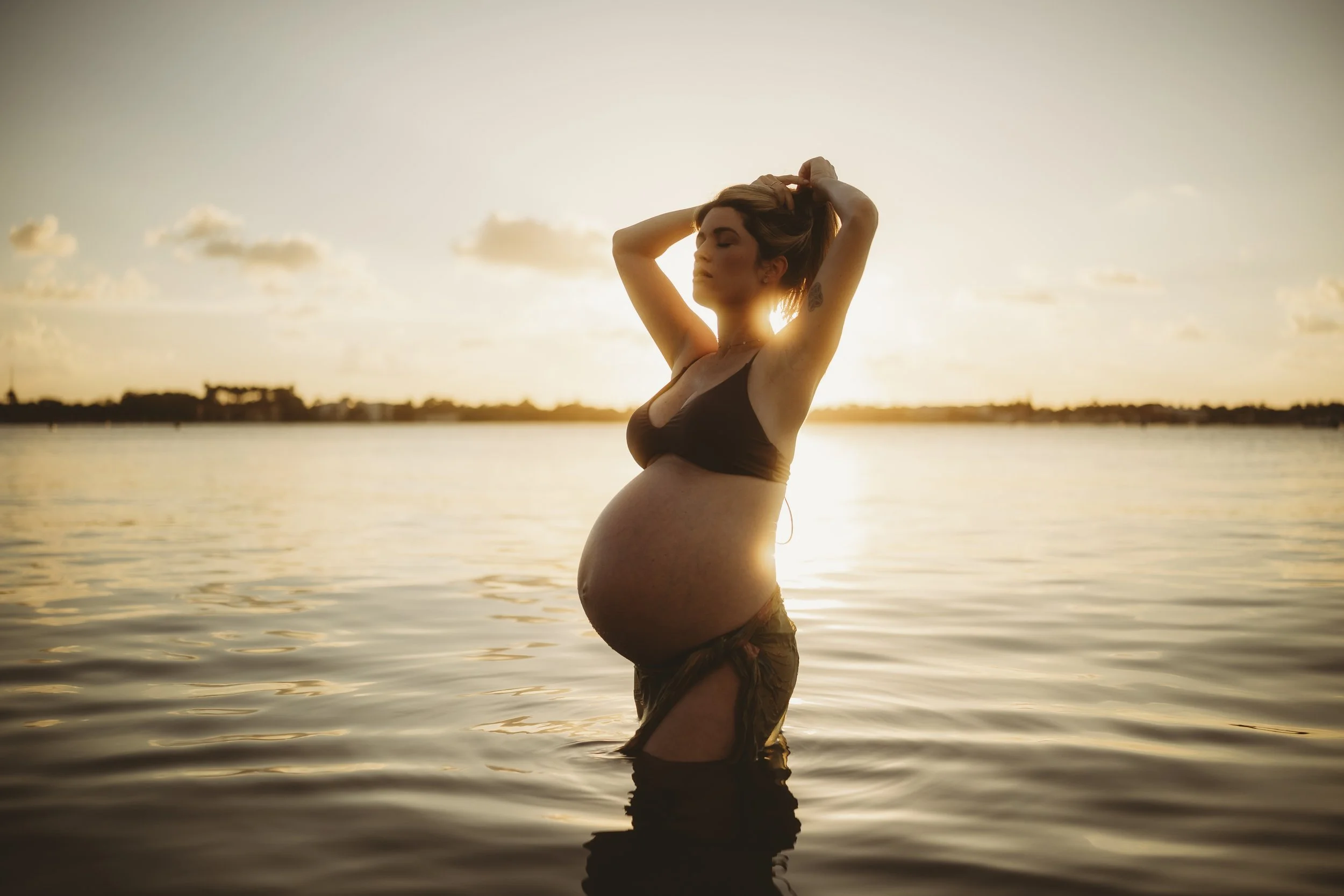 Golden hour maternity portrait of an expectant mother standing waist-deep in calm water, silhouetted by the setting sun as she lifts her hair and embraces the glow of motherhood