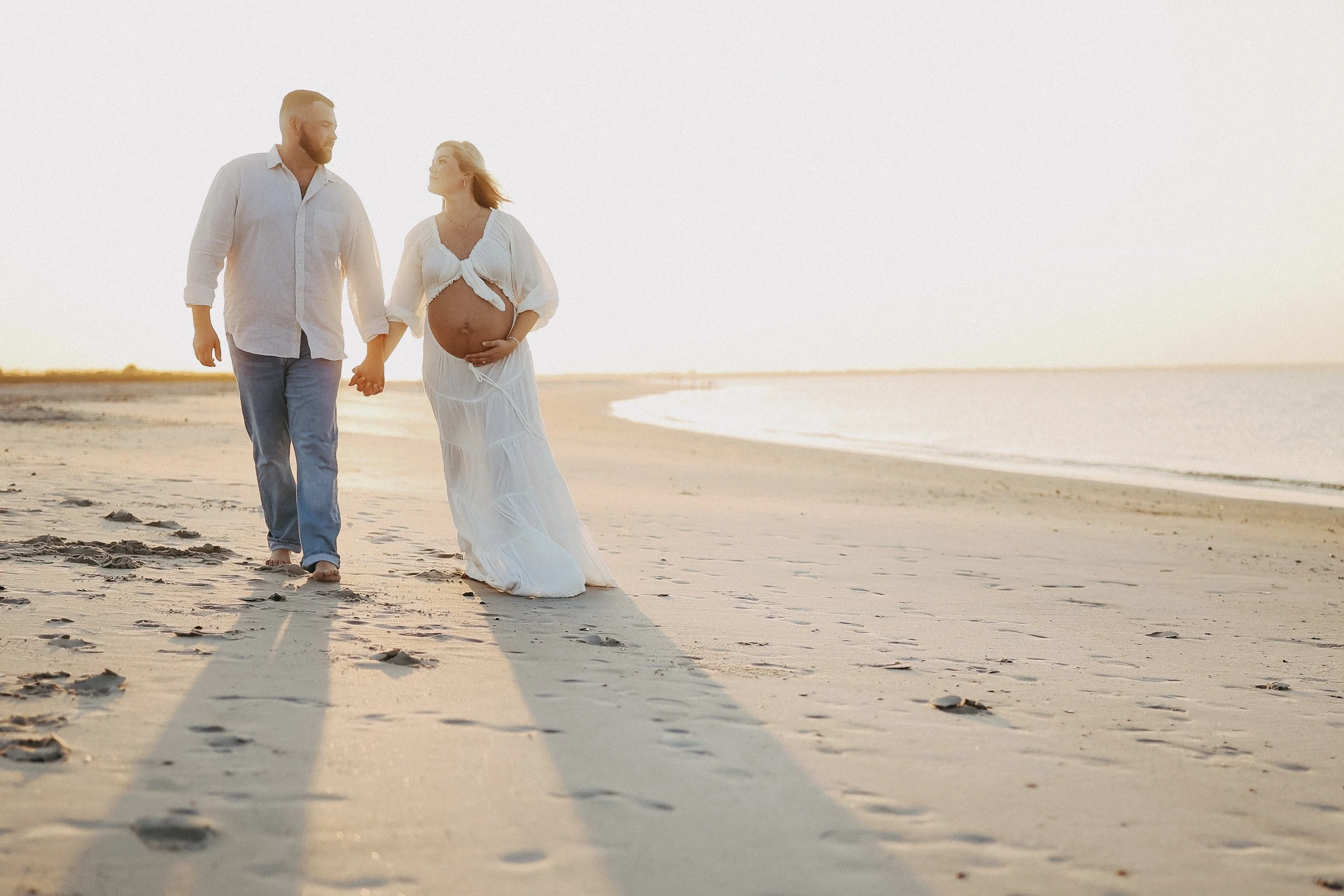 Ethereal sunset maternity image of an expectant couple strolling along the beach, surrounded by soft light and peaceful coastal tones.