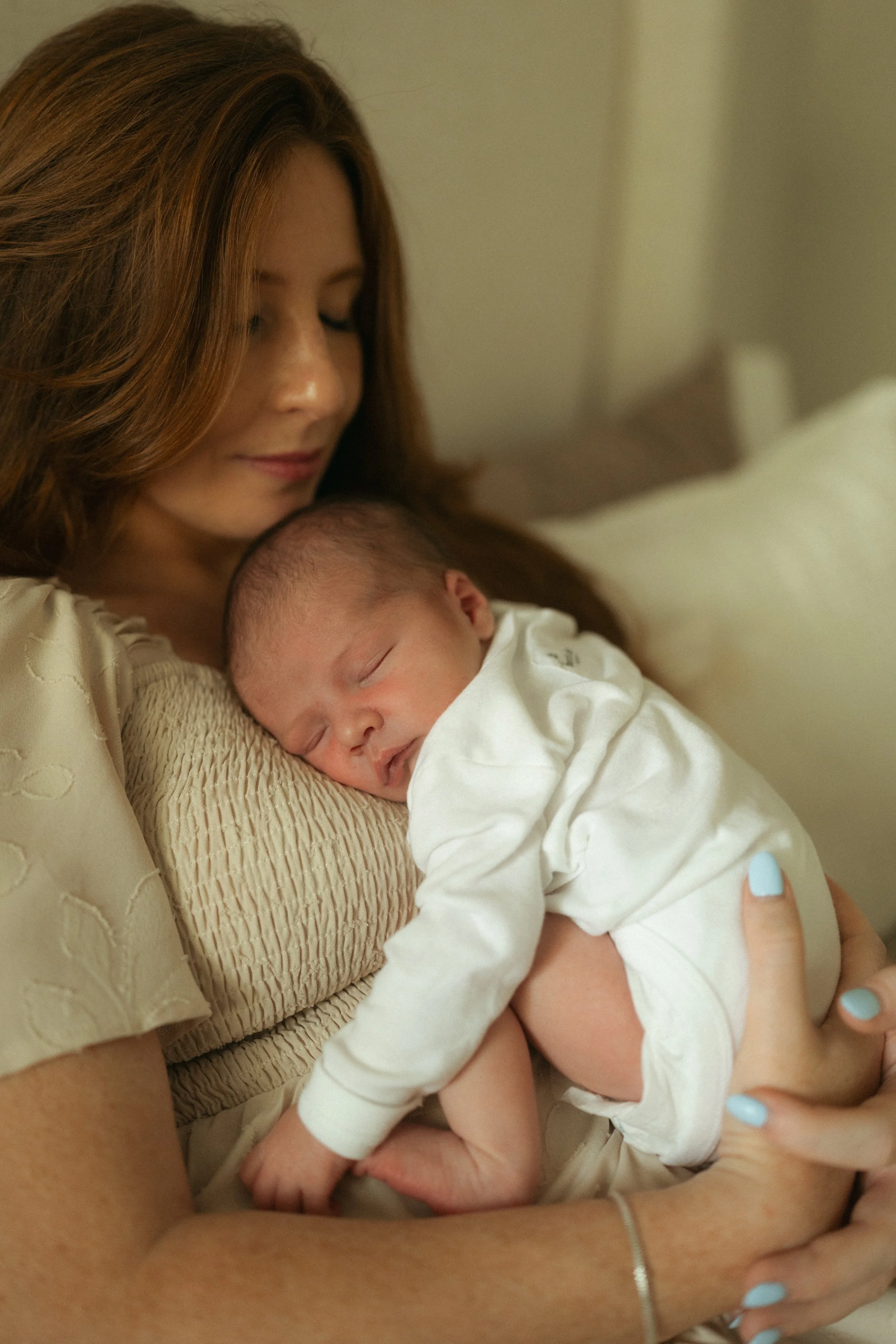Sleeping newborn rests peacefully against their mother's chest as she gently cradles them, creating a quiet, intimate moment during an in-home lifestyle newborn photography session.