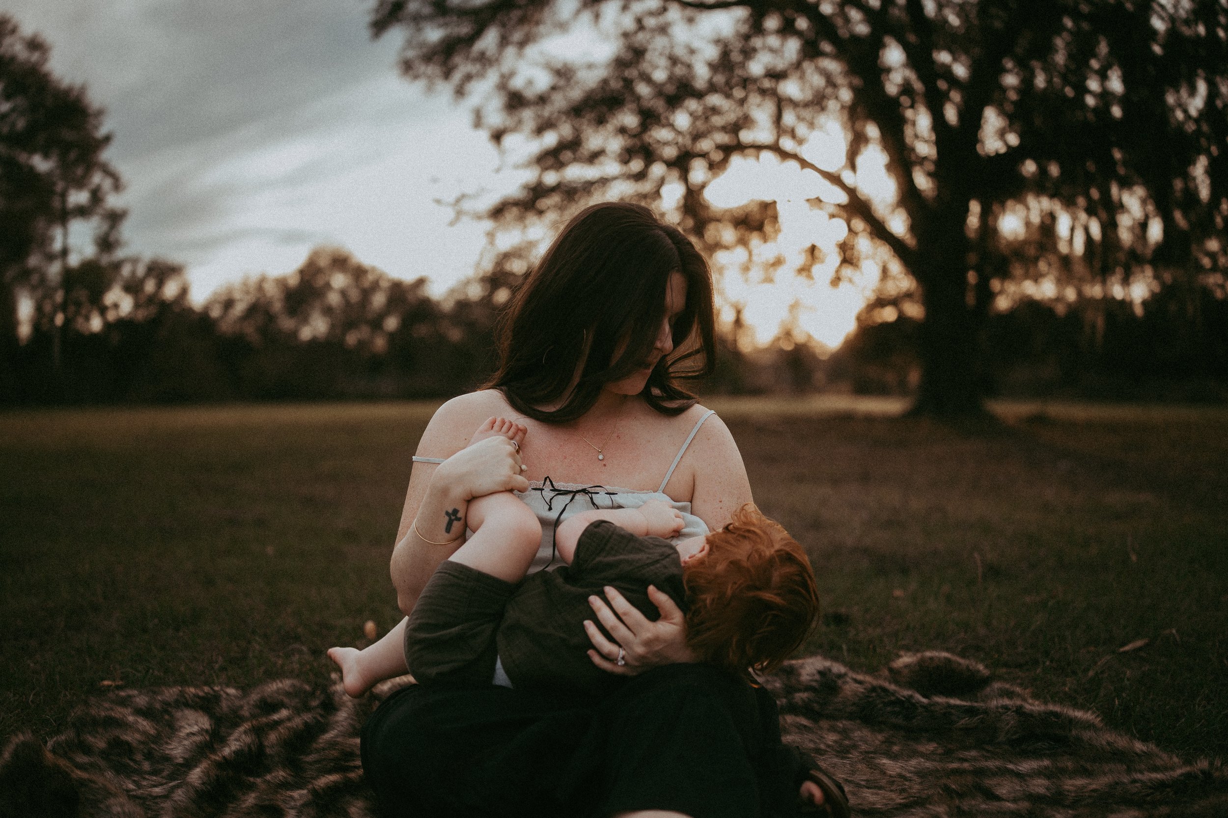A mother breastfeeds her toddler while cradling her baby during a peaceful outdoor motherhood session.