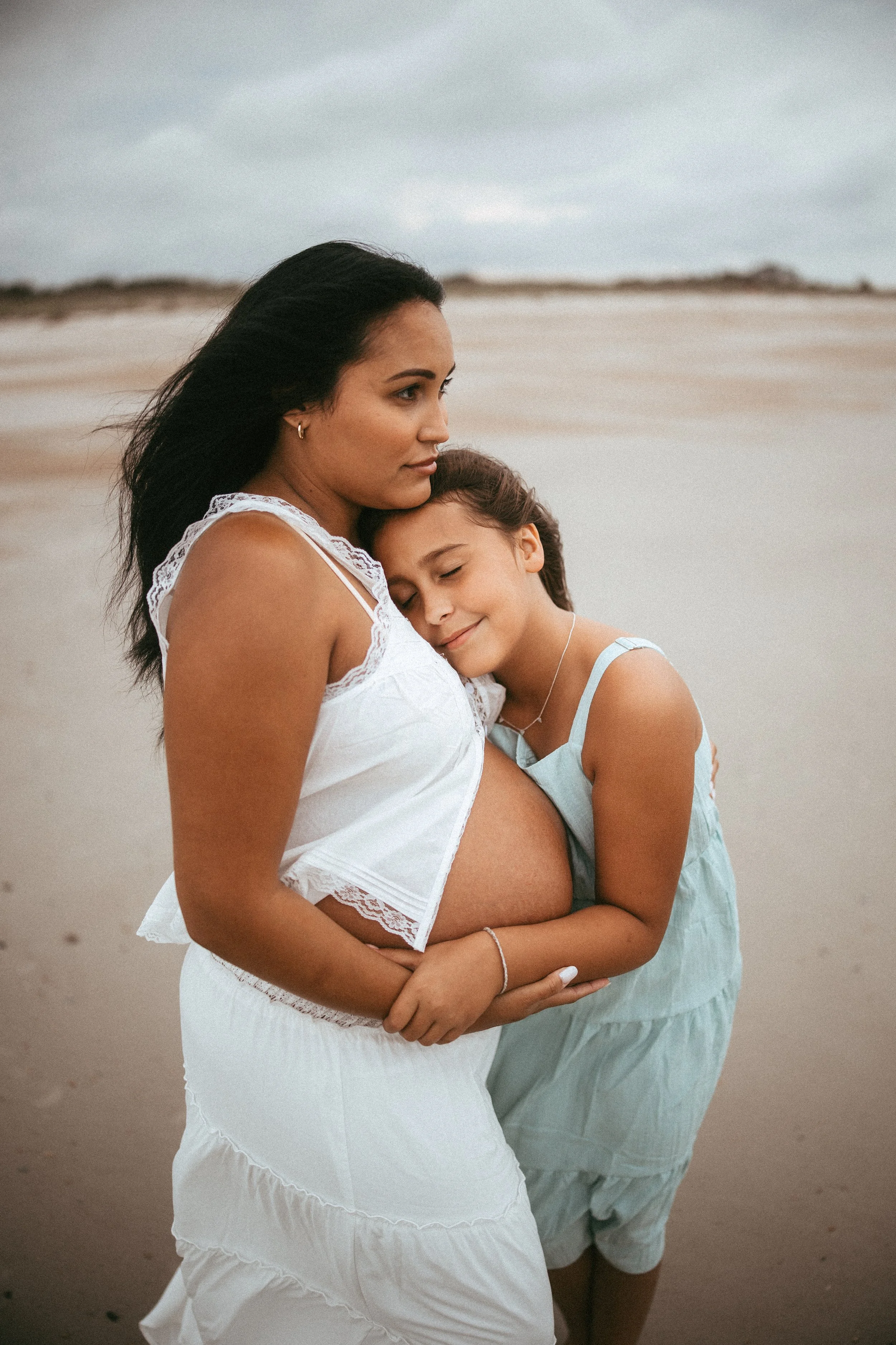 Young girl hugs her mother's pregnant belly on the beach at Fort Clinch State Park in Fernandina Beach, Florida, sharing a sweet sibling moment during a heartfelt maternity photography session.