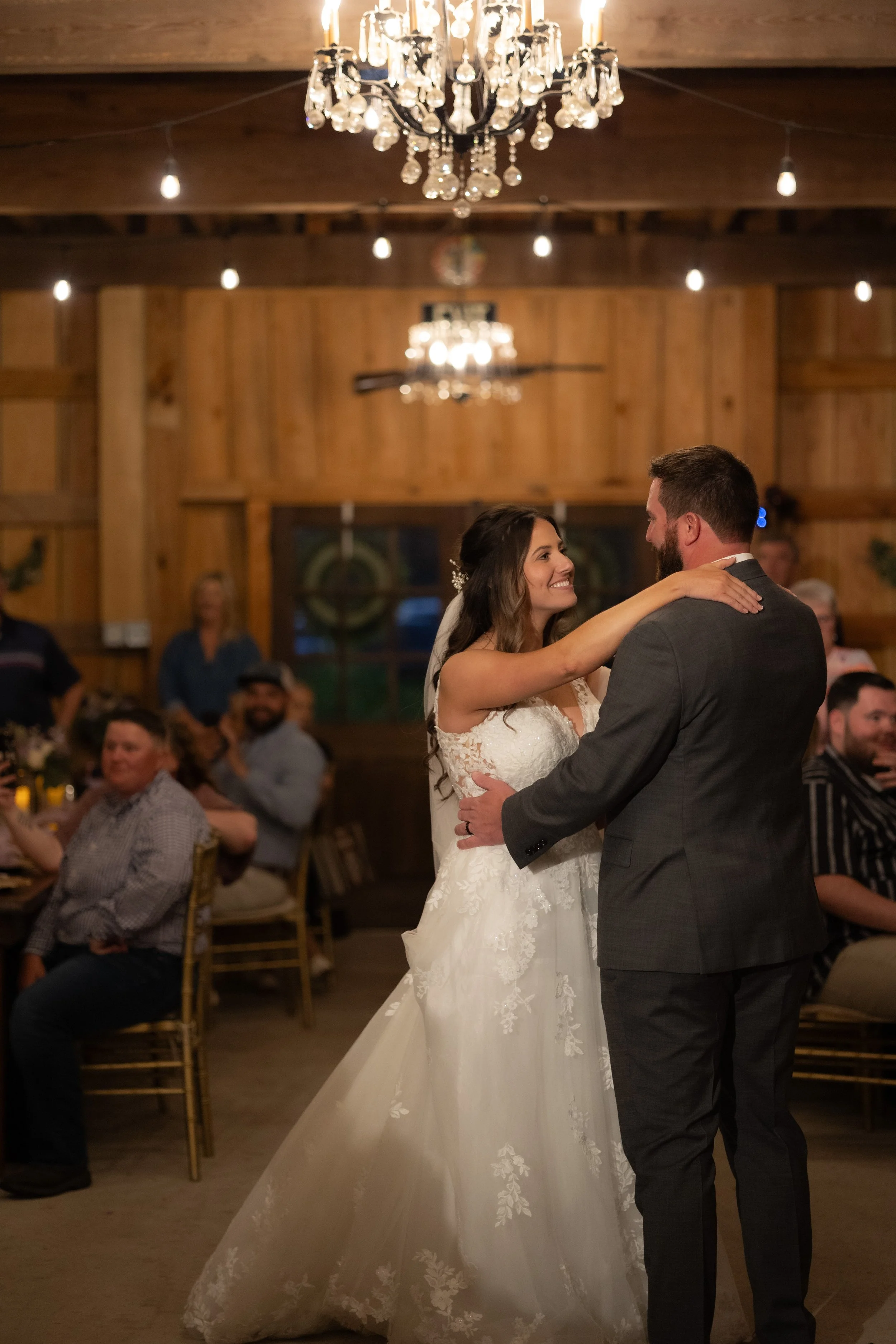 Bride and groom sharing their first dance beneath chandeliers as guests watch during the reception.