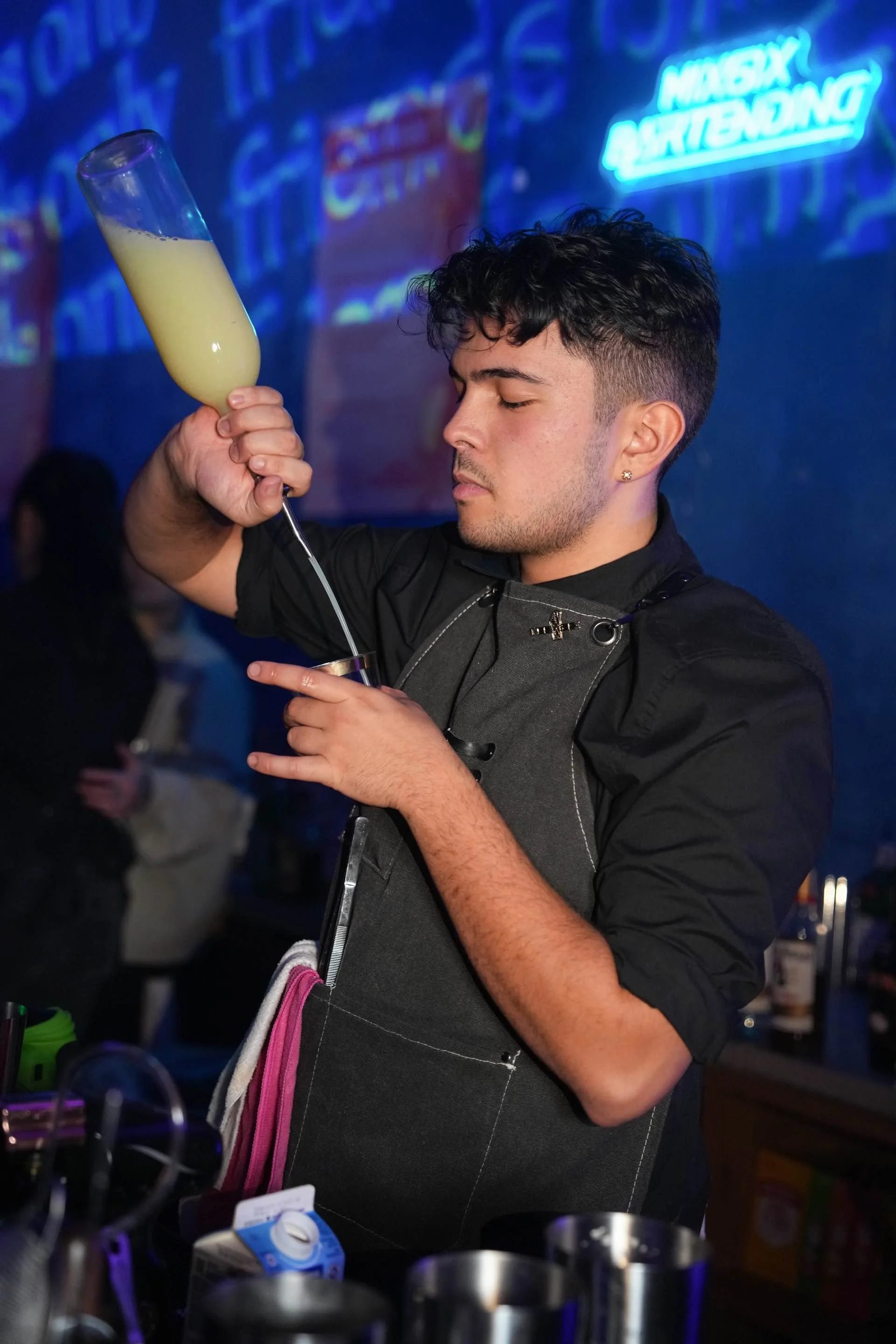 A bartender in black attire and a dark apron pours a yellow cocktail into a shot glass in a bar with neon signs in the background.