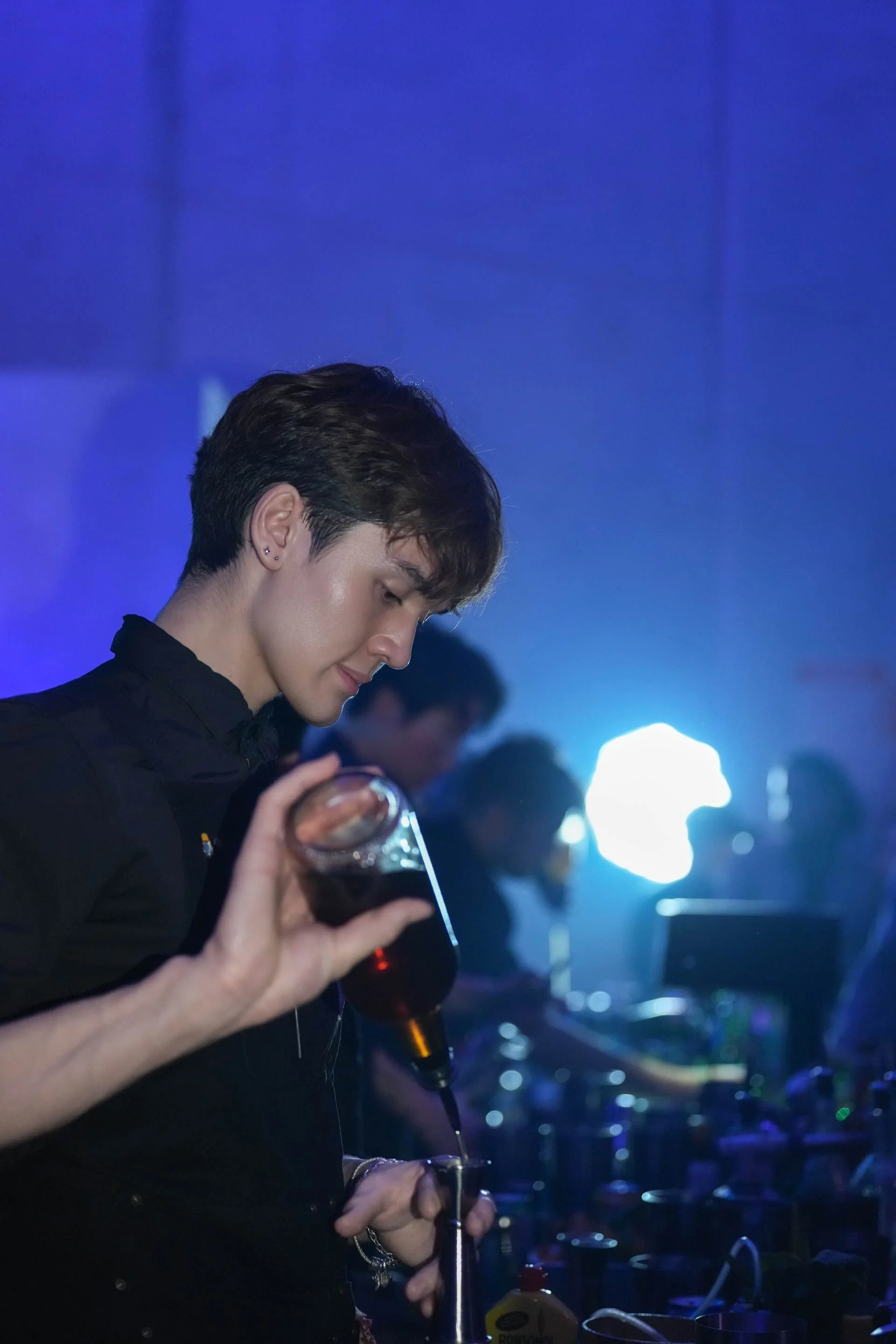 A man with short dark hair and earrings pouring a dark soda from a bottle into a glass at a party or nightclub with blue and purple lighting, with other people in the background.
