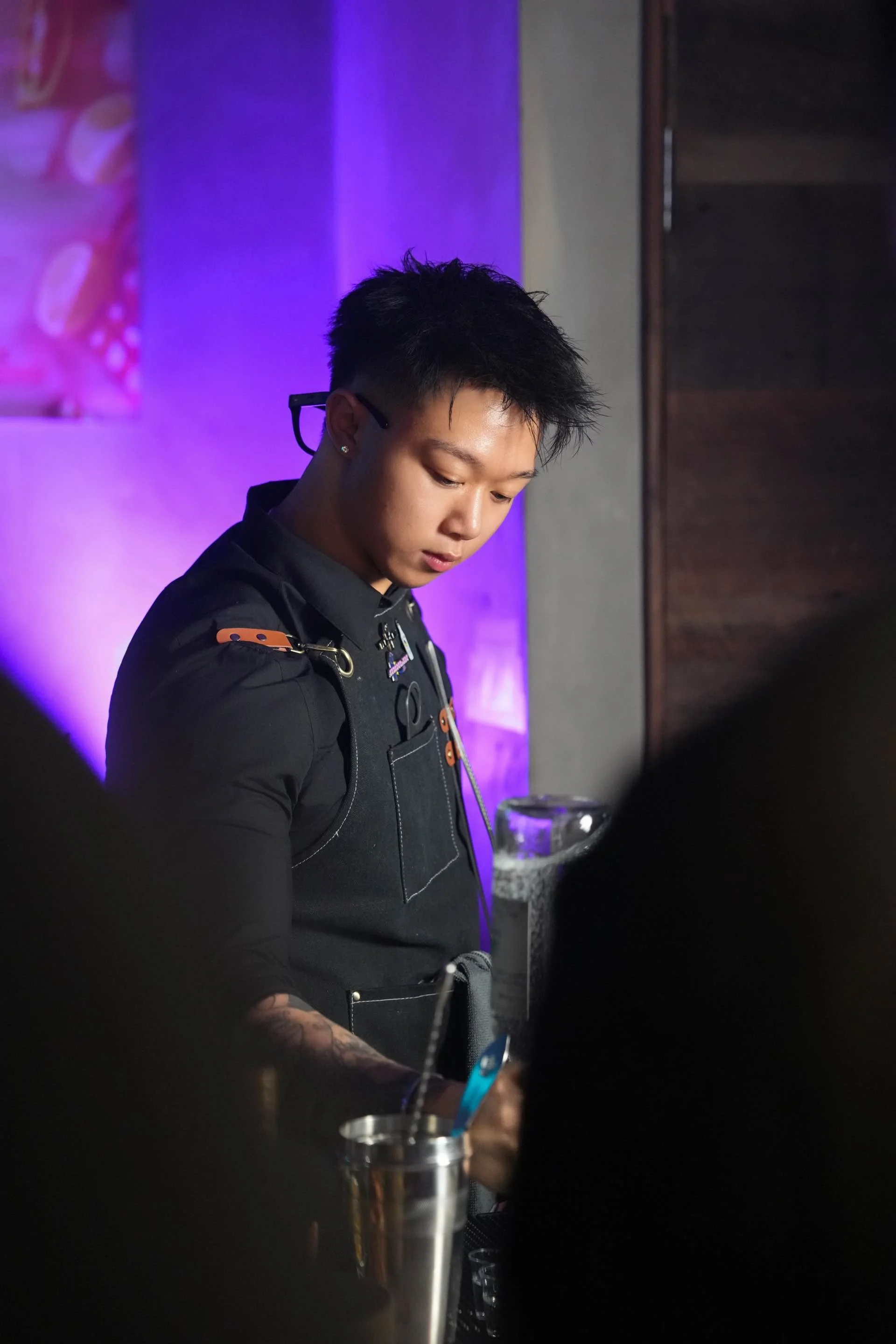 A young man with short black hair, wearing glasses and a black outfit, standing at a bar with a purple backdrop, focused on preparing a drink.