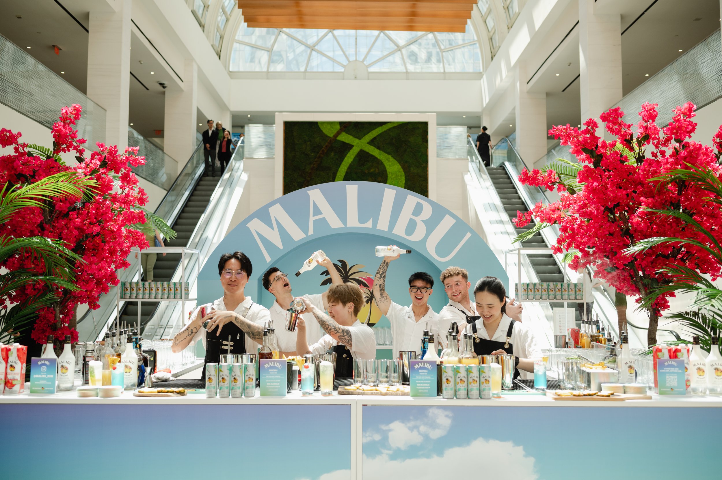 Group of six people behind a Malibu-themed bar at an indoor mall, with pink flowering trees on either side, and escalators in the background.