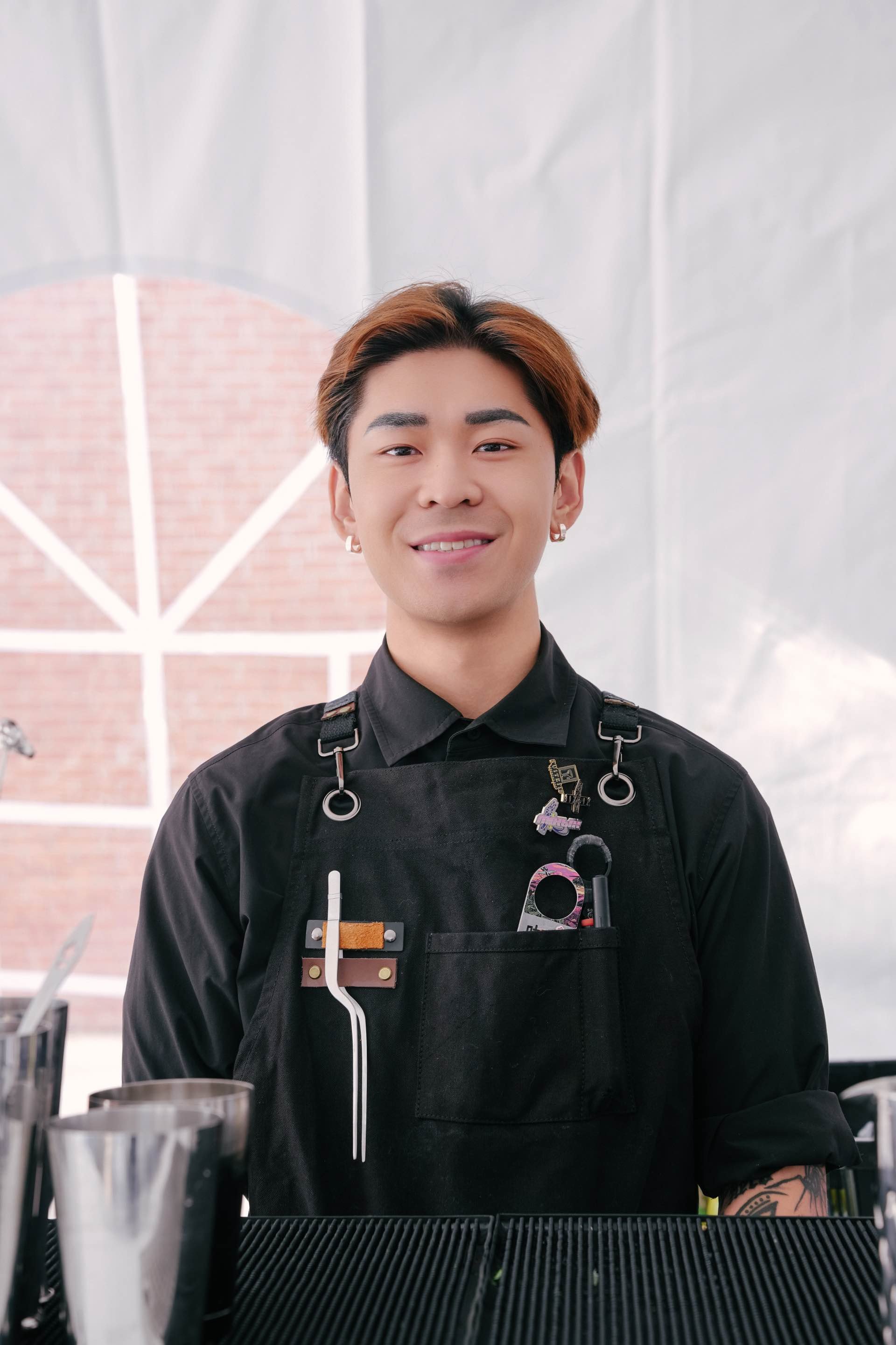 Young man with tattoos and earrings smiling, wearing a black apron with bar tools, standing behind a bar counter with glasses, in front of a window with brick building outside.