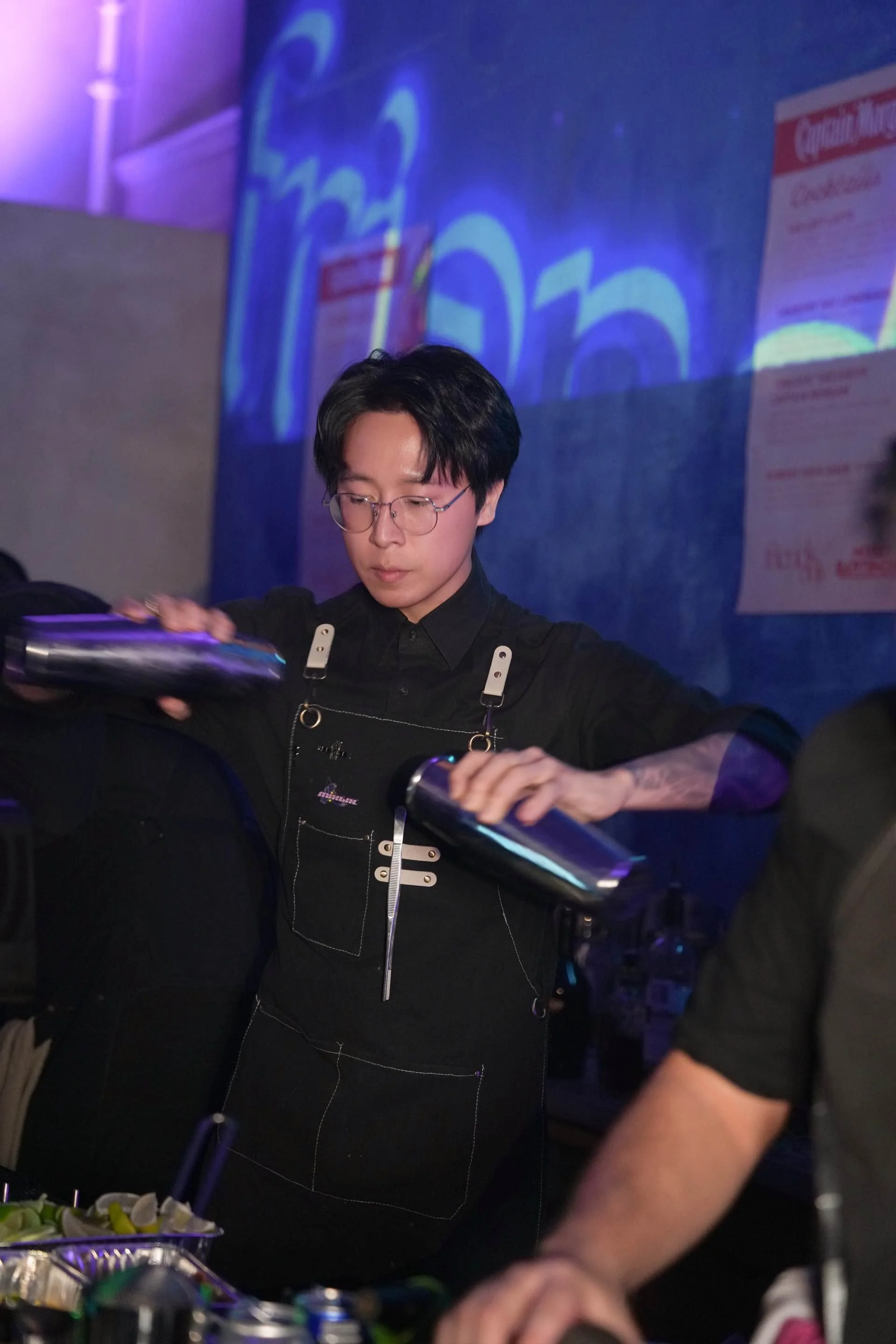 A young man wearing glasses and a black apron prepares drinks behind a bar in a dimly lit setting with blue and purple lighting.