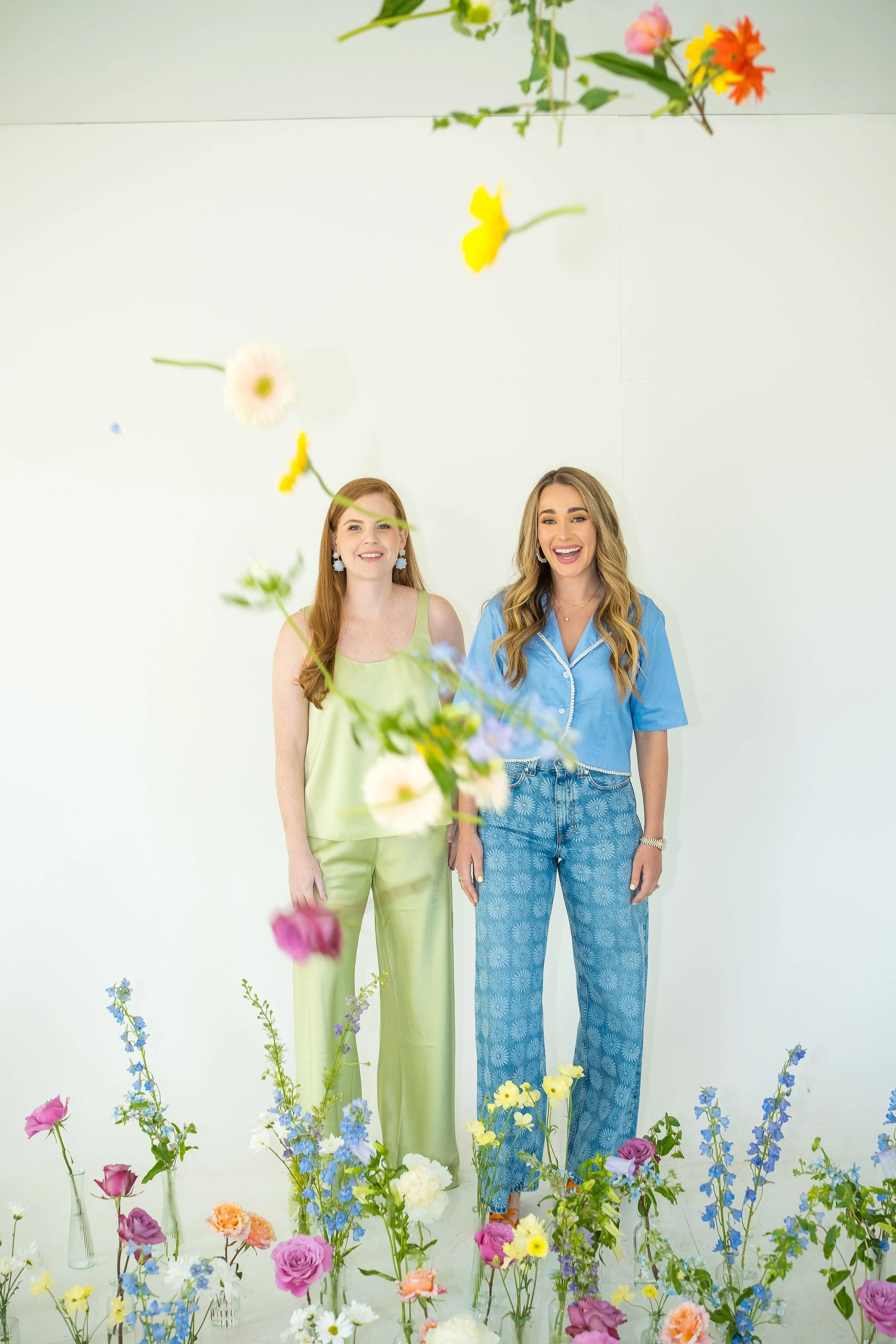 Two women standing surrounded by colorful flowers in vases, smiling, with a backdrop of a white wall decorated with hanging flowers.