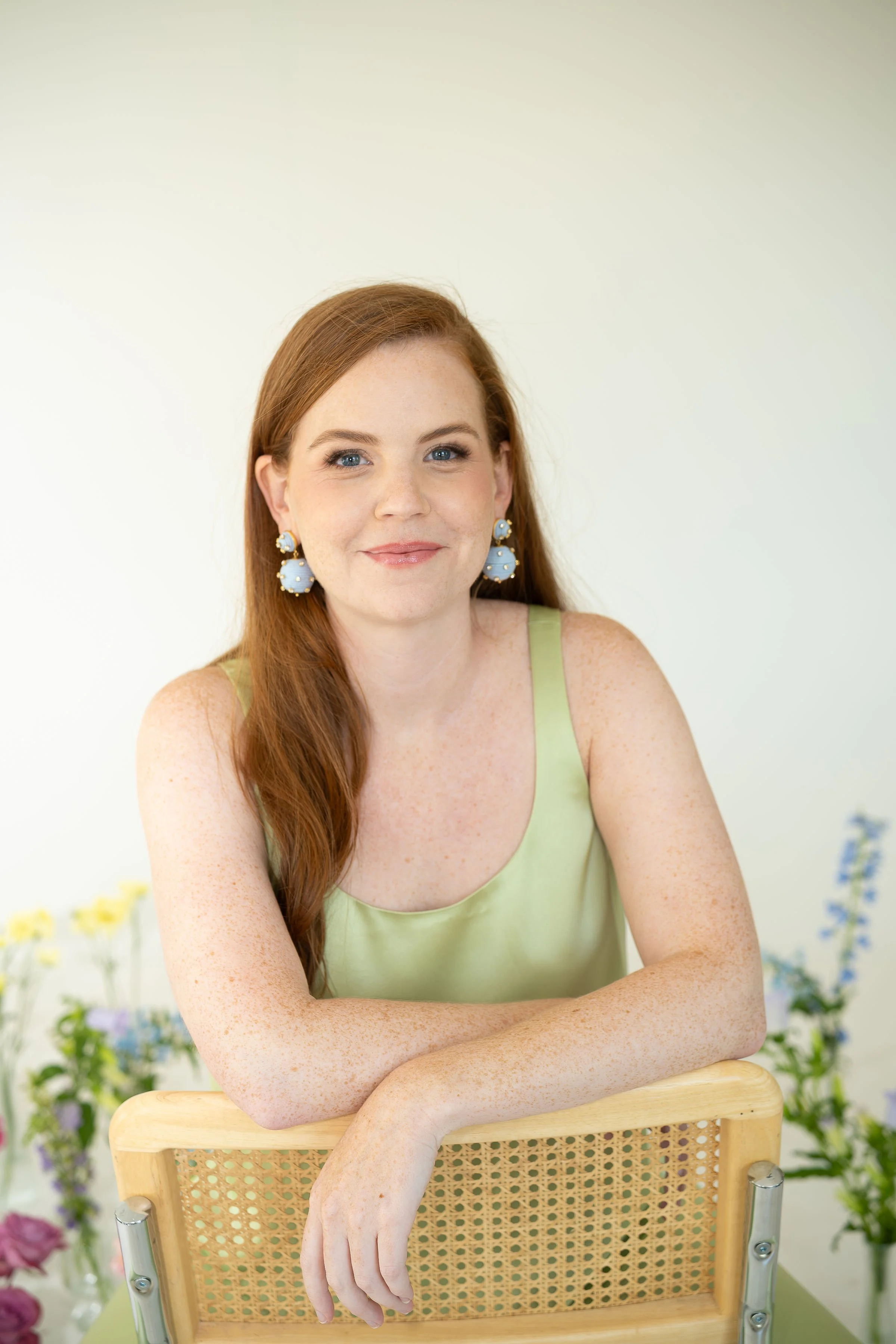 A young woman with red hair and blue eyes, wearing a light green sleeveless top and large blue earrings, is sitting at a wooden table with her arms resting on a wooden chair, smiling softly with a floral background.