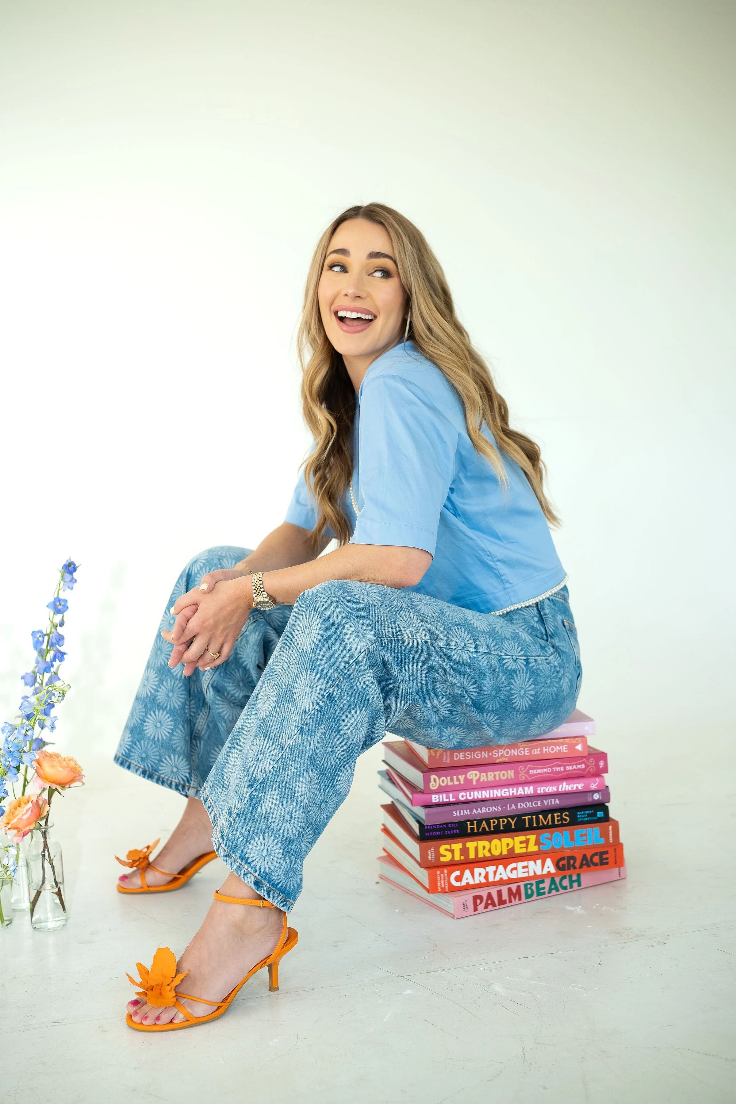 A woman with long wavy hair, wearing a blue crop top and wide-leg patterned jeans, sitting on a stack of colorful books, wearing orange high heels with floral embellishments, smiling and looking to the side. There are flowers in glass bottles on the floor, and a plain light green background.