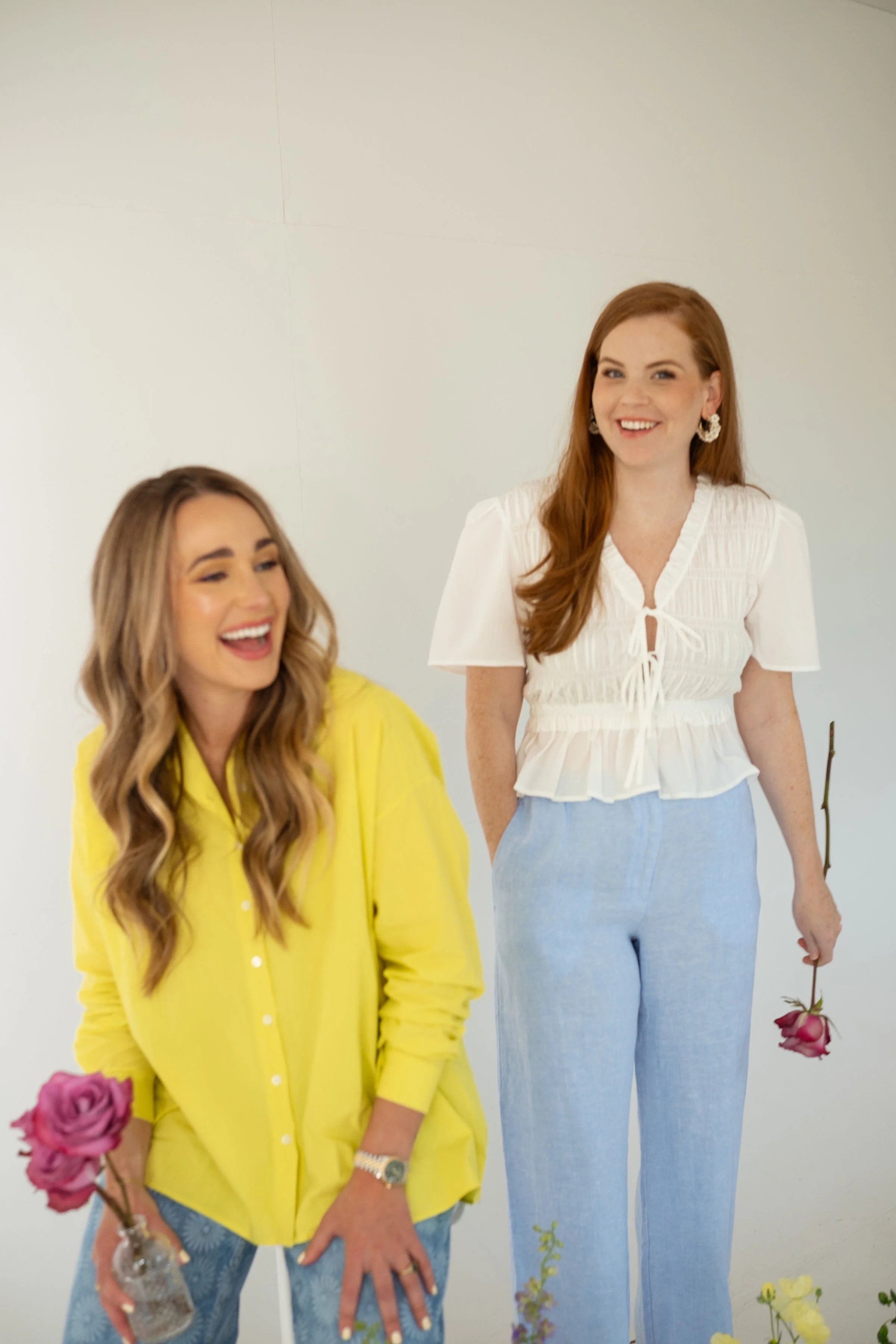 Two women with long hair, smiling, standing against a plain background. The woman on the left has blonde hair and is wearing a yellow shirt and jewelry, and holding a pink rose in a glass vase. The woman on the right has red hair, wearing a white blouse and light blue jeans, holding a pink rose at her side.