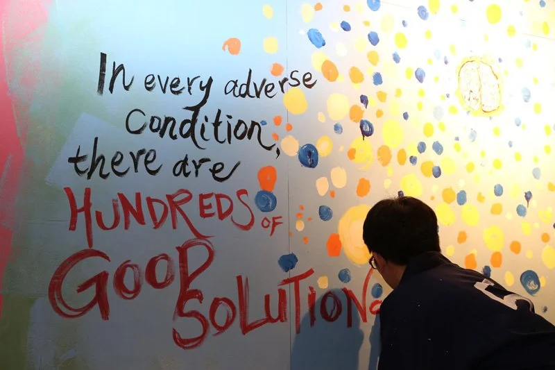 A person with dark hair and glasses looking at a colorful wall with painted dots and a handwritten quote that says, "In every adverse condition, there are hundreds of good solutions."