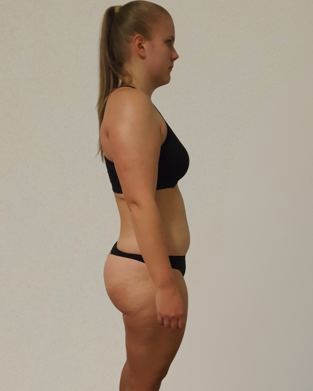 Side view of a woman with fair skin in black underwear, standing against a plain off-white wall.