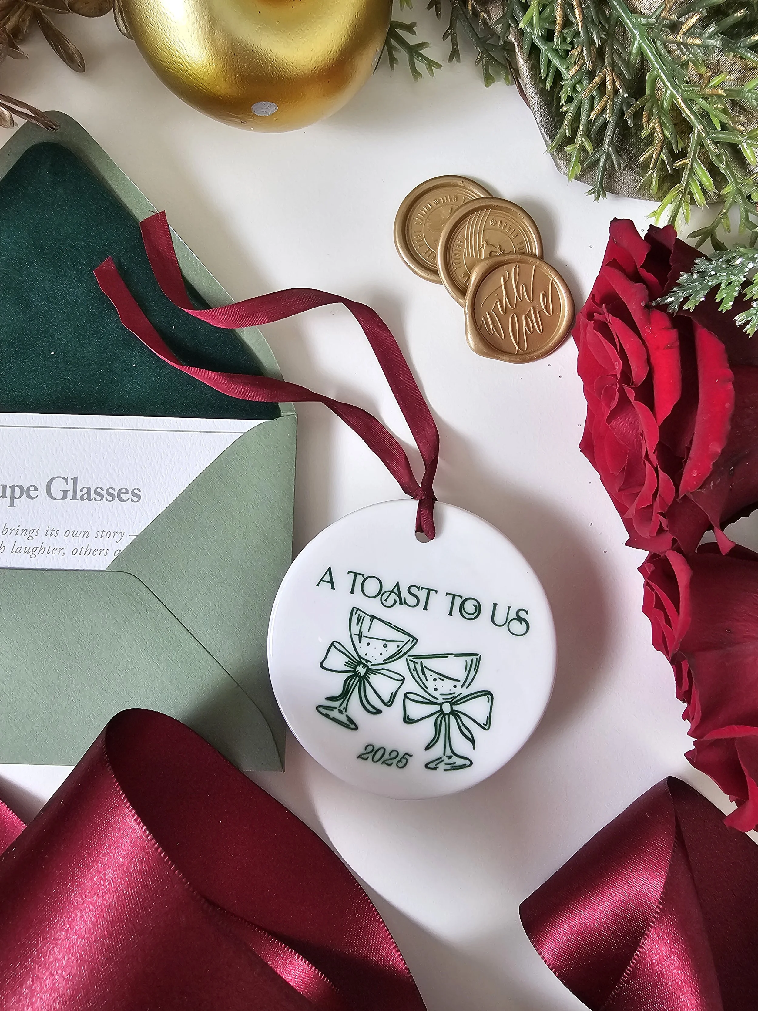A Toast to Us ceramic ornament styled with gold wax seals, greenery, and burgundy ribbon