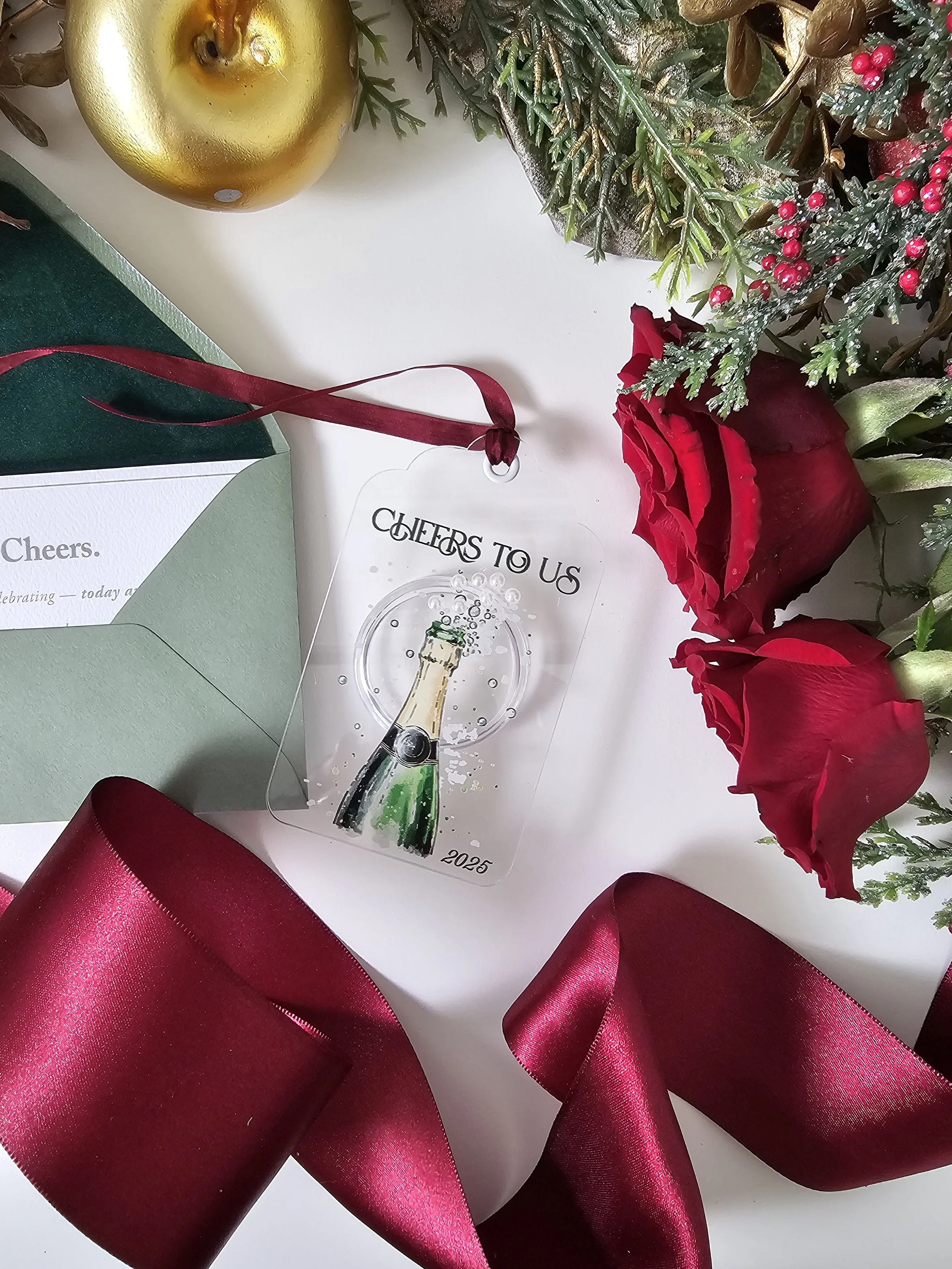 Romantic champagne ornament styled with roses, greenery, gold holiday decor, and red satin ribbon by RBW Stationery and Events, LLC