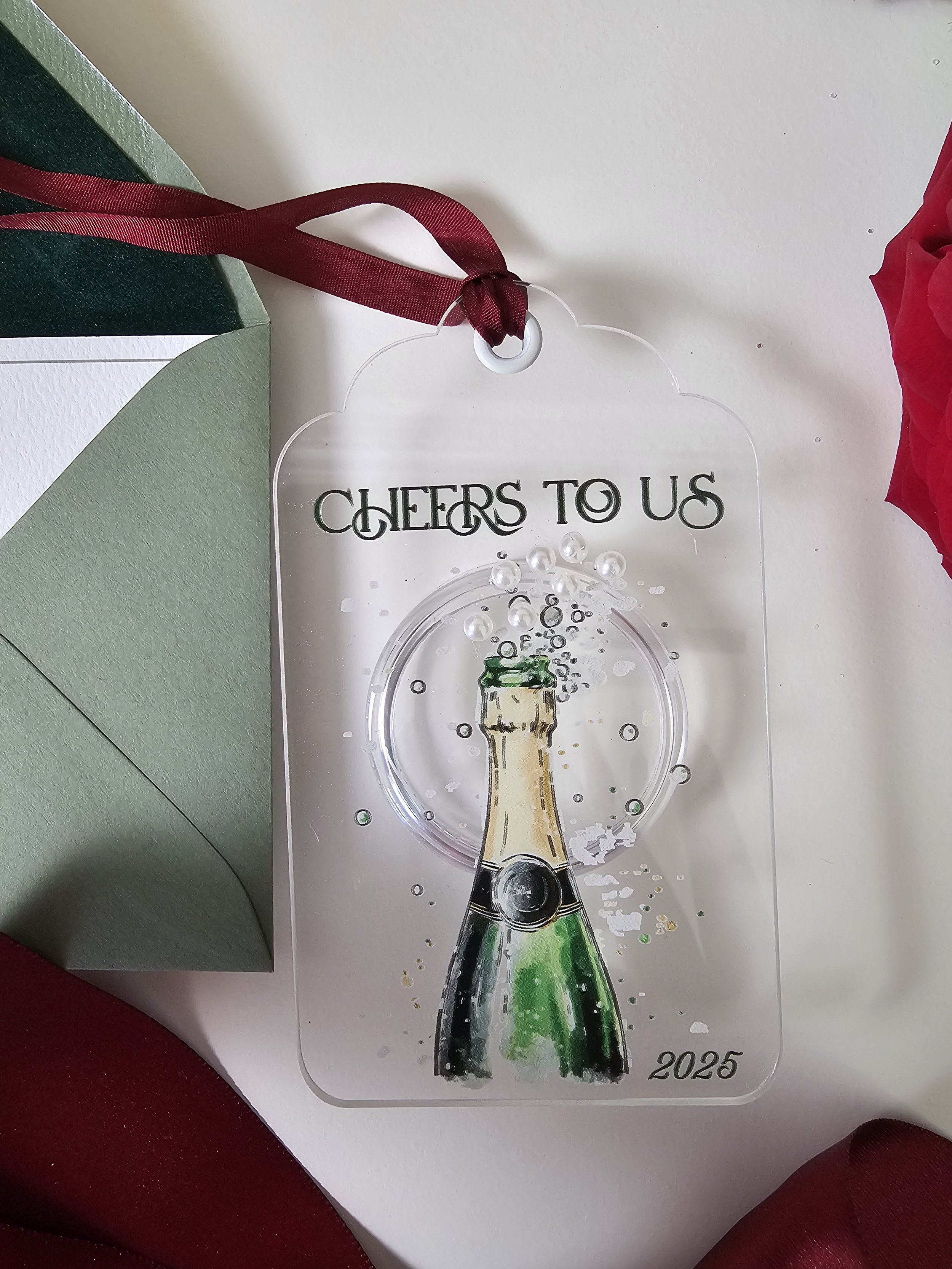 Cheers to Us 2025 champagne bottle ornament with pearl bubble accents and burgundy ribbon by RBW Stationery and Events, LLC