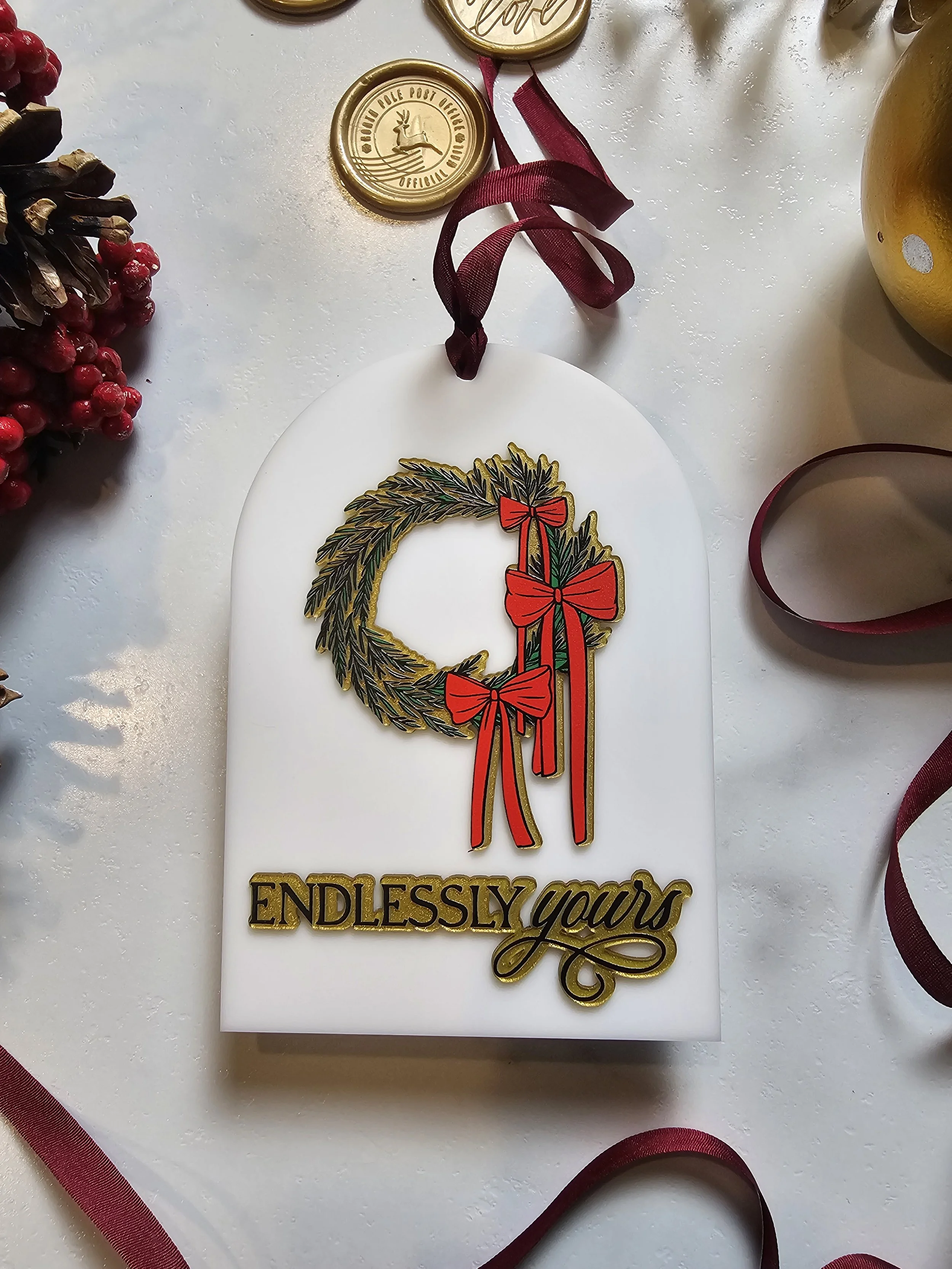 White acrylic Christmas ornament with evergreen wreath and burgundy silk ribbon displayed with holiday accents