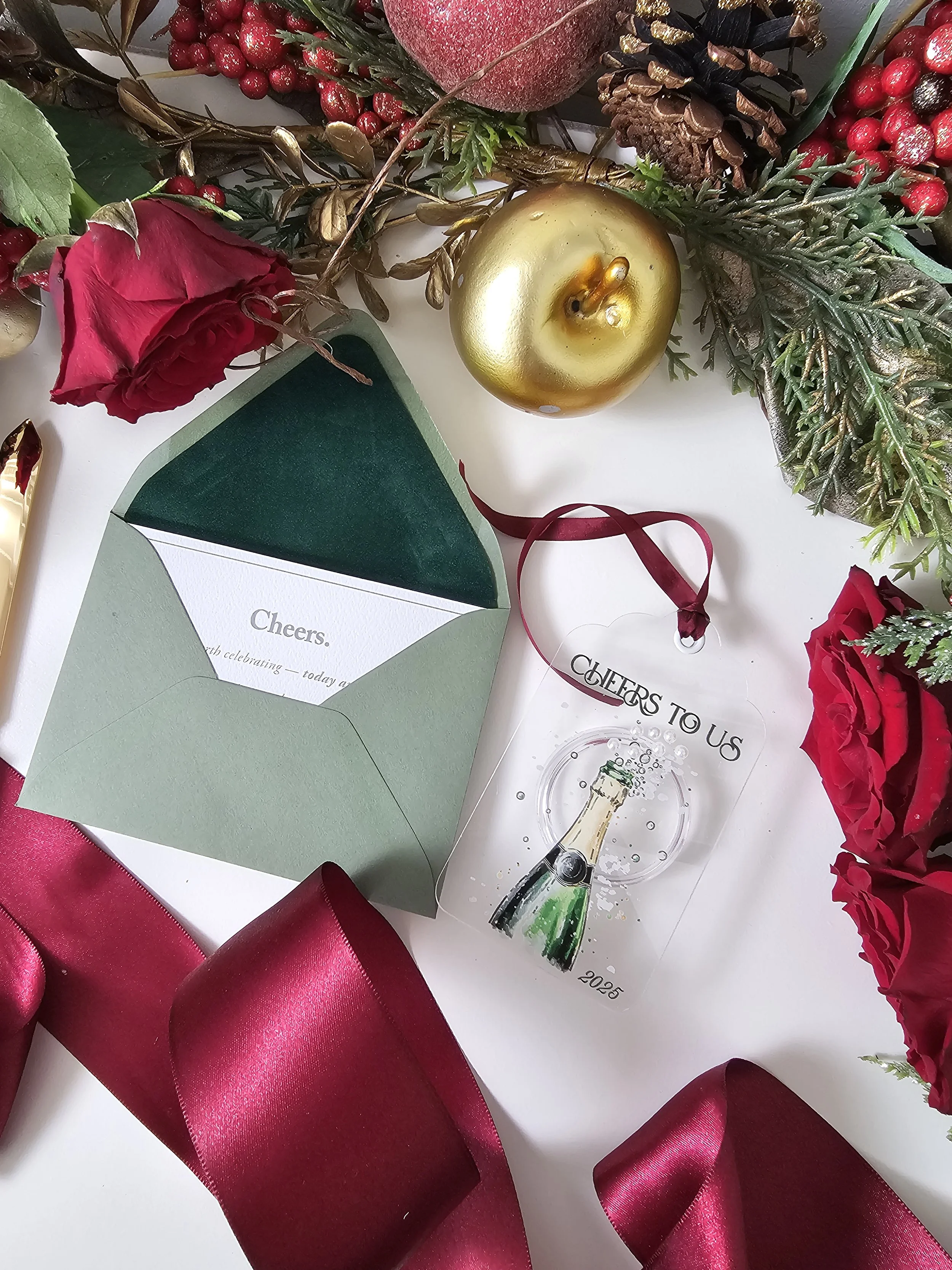 Flatlay of champagne ornament with deep red roses, burgundy ribbon, and green velvet envelope by RBW Stationery and Events, LLC