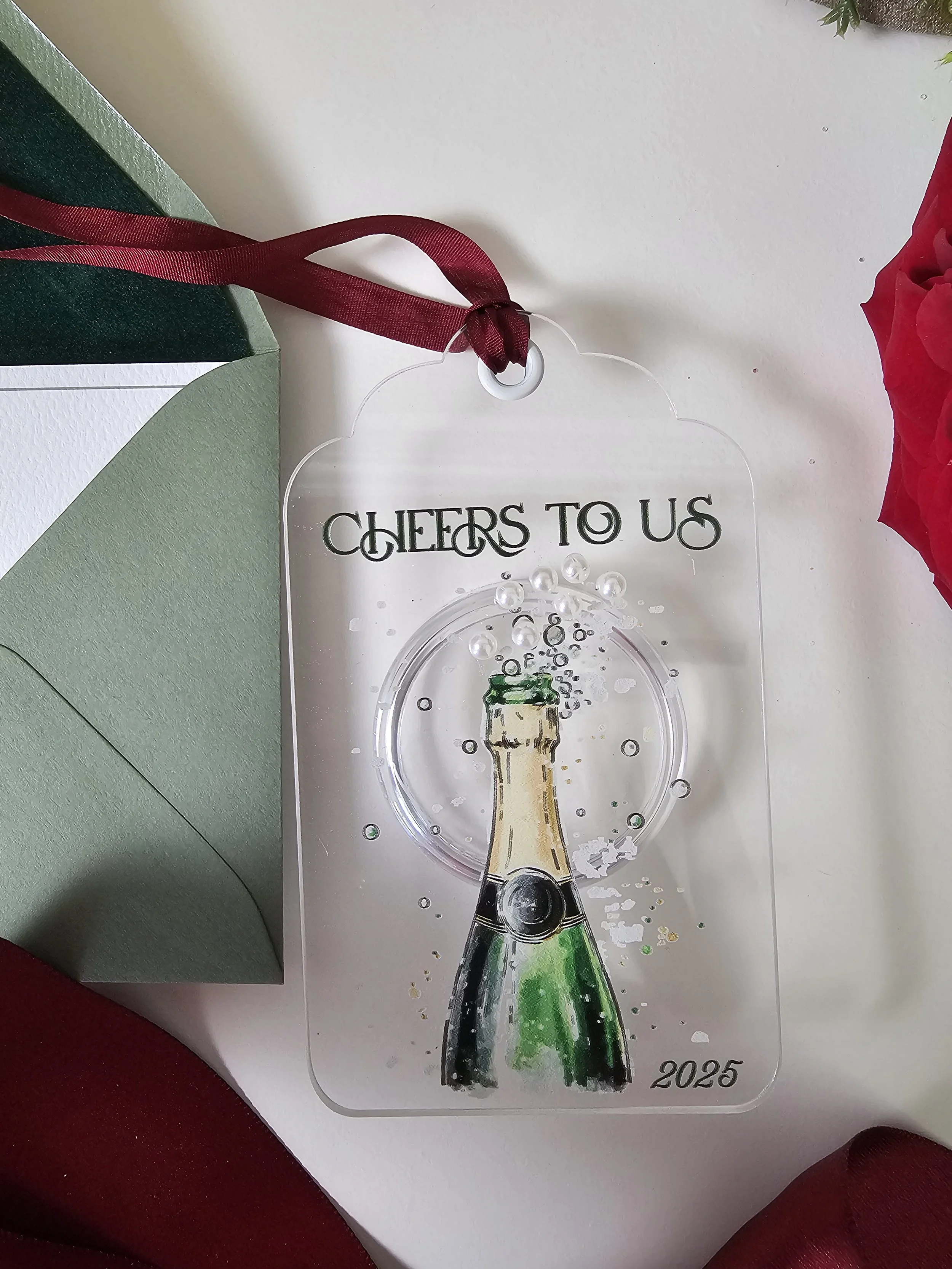 Close-up of the ‘CHEERS TO US’ 2025 ornament showing champagne detail and burgundy ribbon by RBW Stationery and Events, LLC