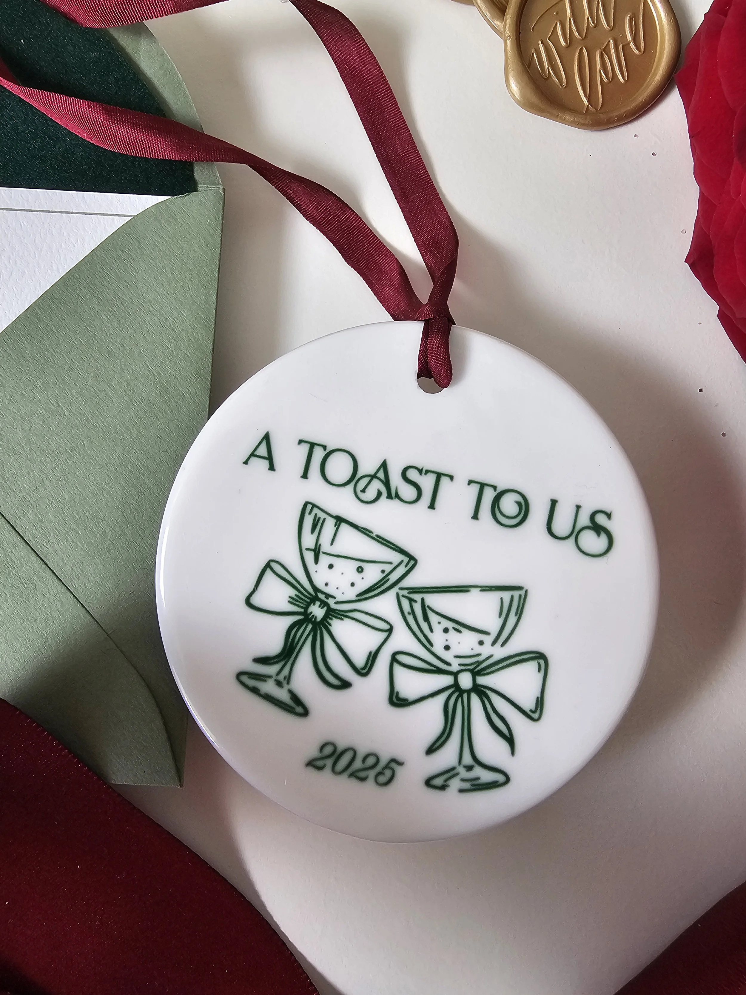 Seasonal flatlay with A Toast to Us ceramic ornament, burgundy ribbon, and festive decor