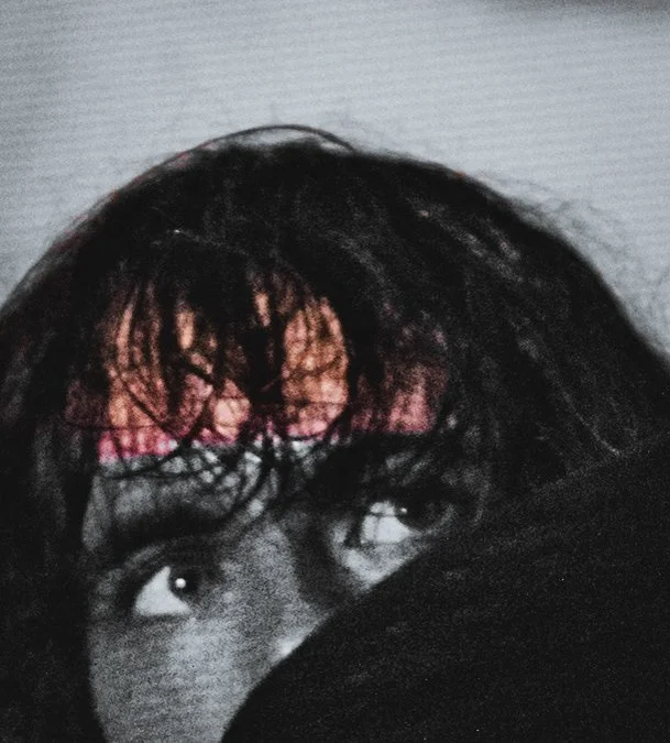 Close-up photo of a person's face with dark, tousled hair, looking upward, partially obscured by a drum cymbal