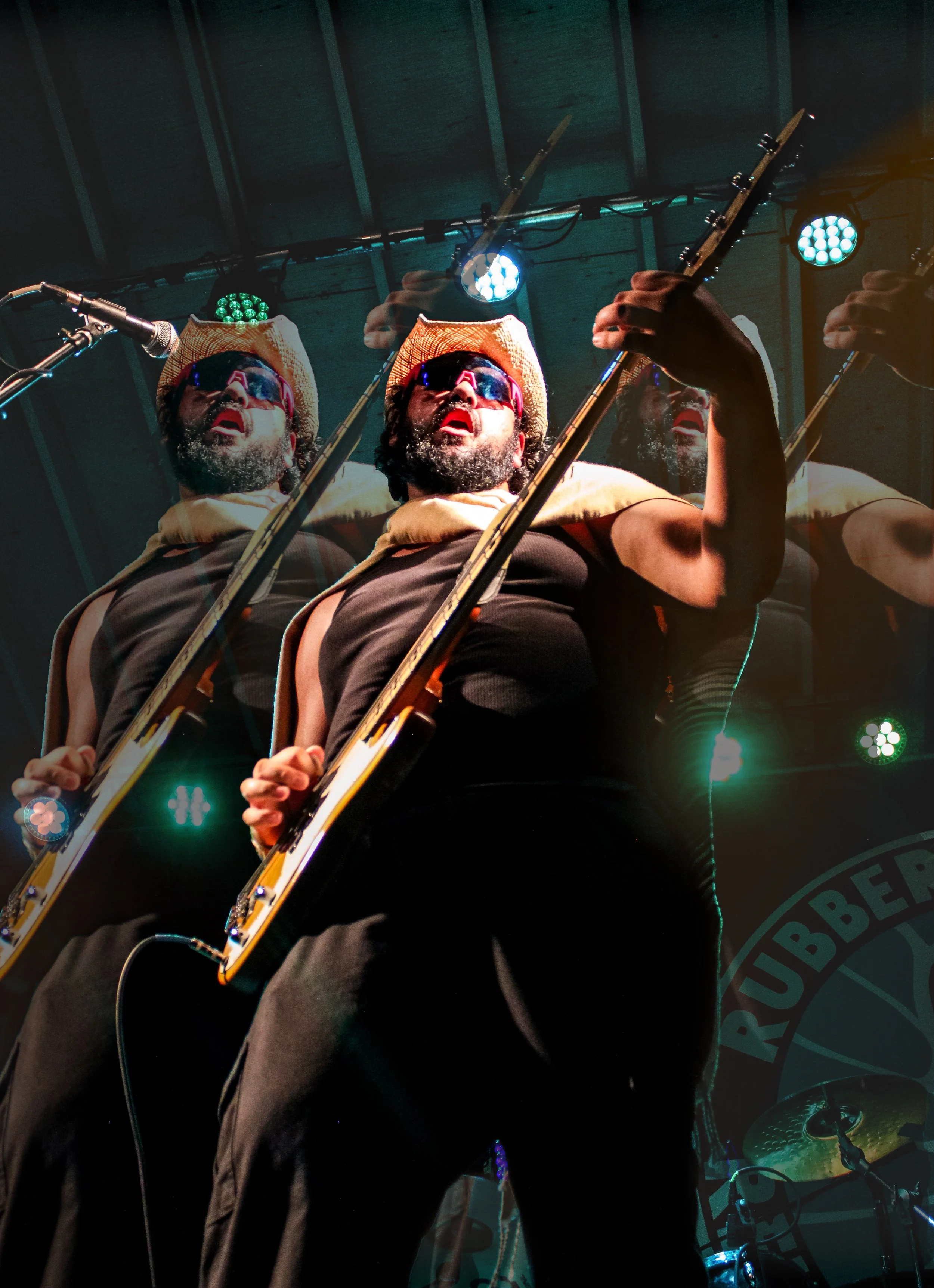A musician wearing a straw hat and sunglasses playing a guitar on stage, with stage lights overhead and a drum set in the background.