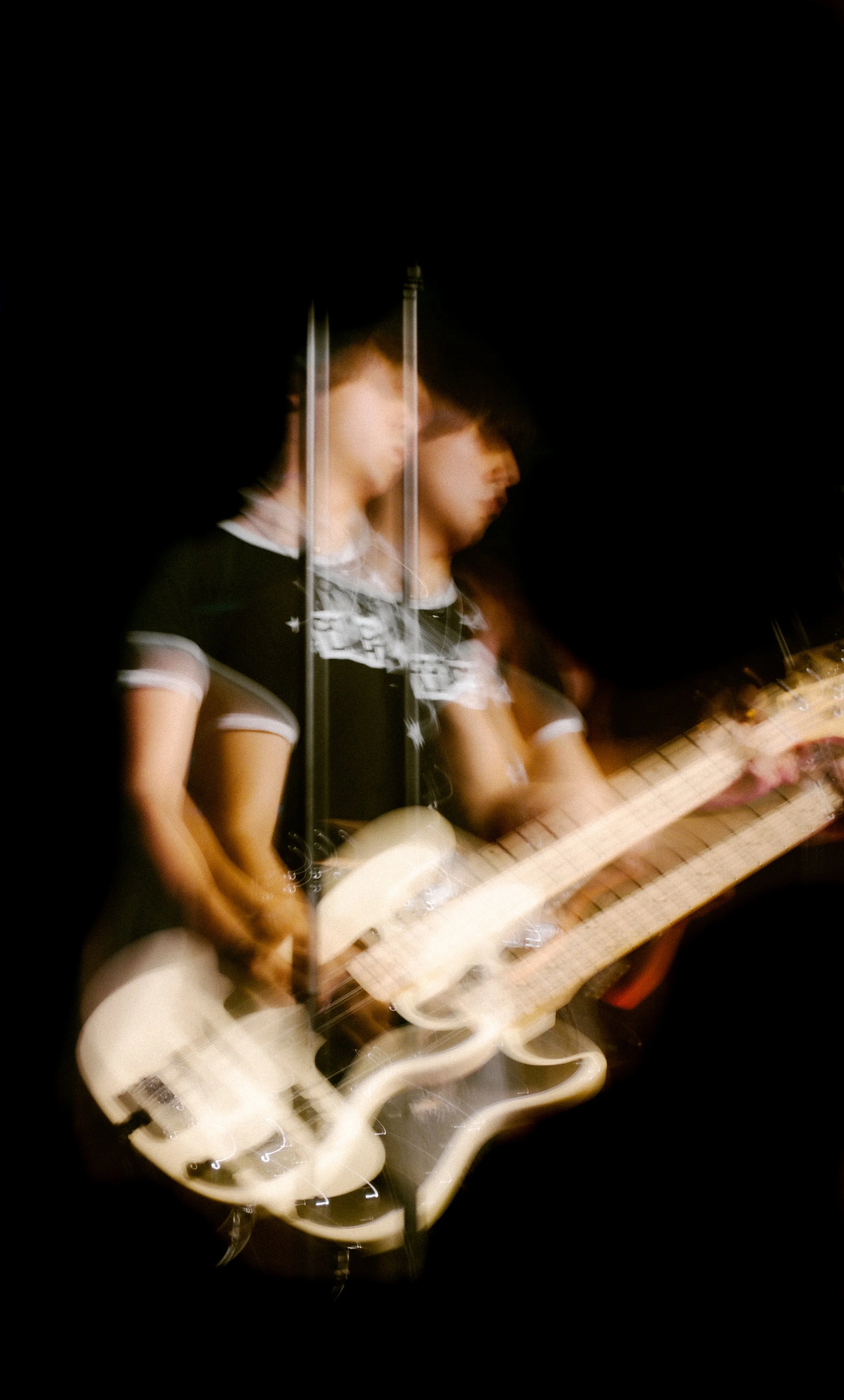 A man with dark hair wearing a black t-shirt with writings on the front, is playing electric guitar on stage under black lighting, creating a blurred motion effect.