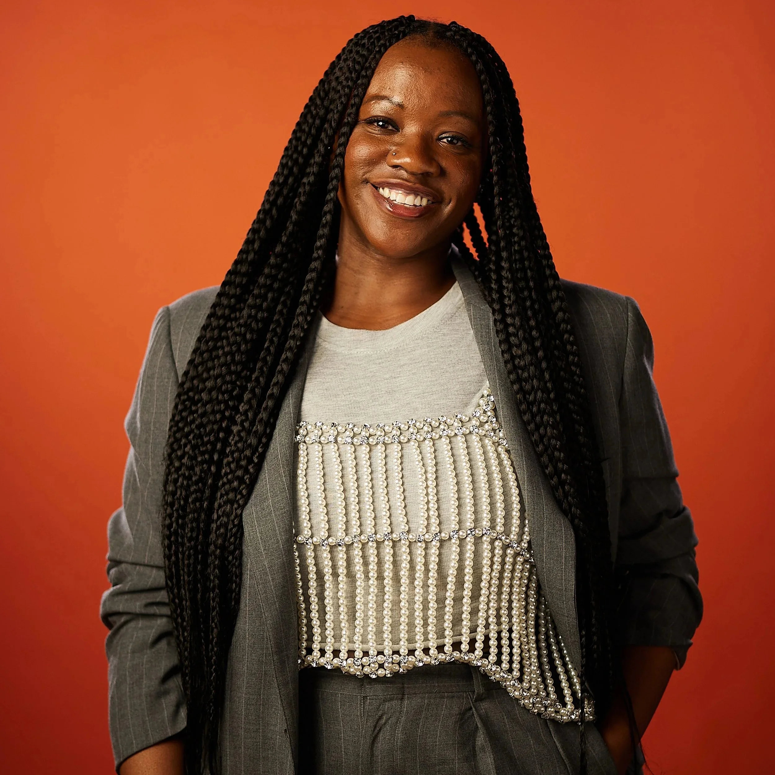 A woman with long braided hair smiling, wearing a grey blazer over a pearl-trimmed top, against an orange background.