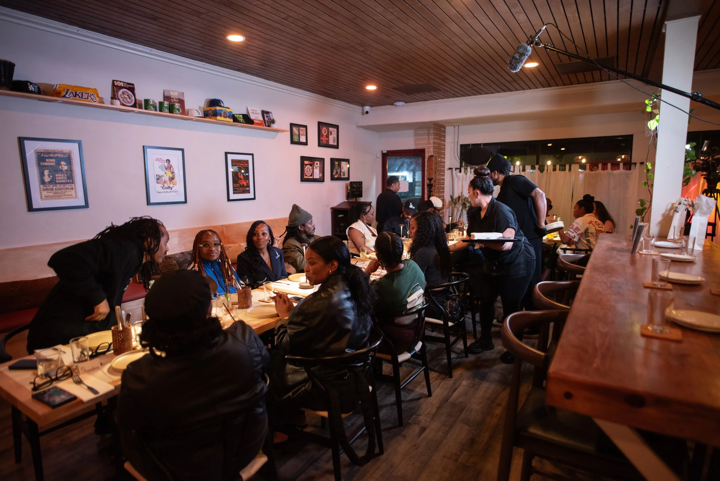 A group of people dining at a restaurant, with staff serving, framed posters on the wall, and a wooden ceiling.