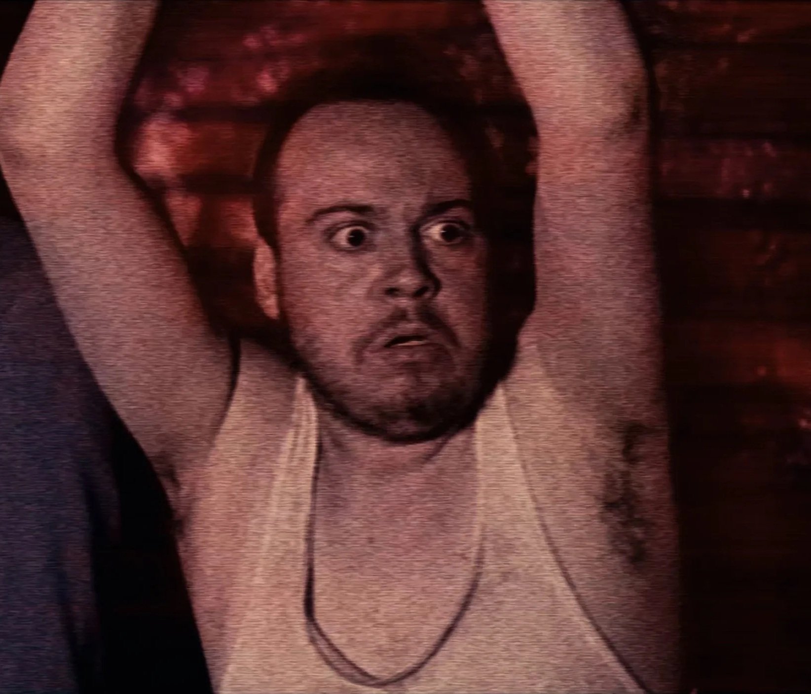 A startled man, wearing a dirty tank top, with their arms restrained their head. Alan Lanxton