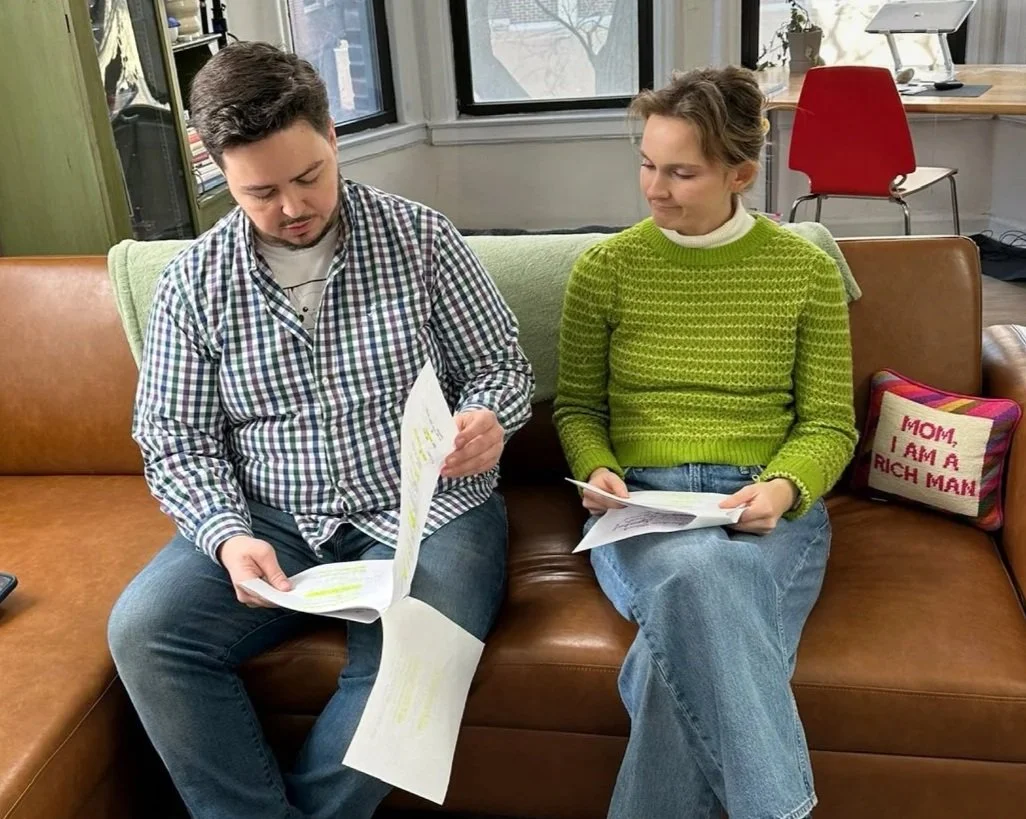 A young man with dark hair in a plaid shirt and a young woman with blonde hair in a green sweater sit on a brown leather couch, reviewing a script. There is a green blanket on the back of the couch and a decorative pillow. Jordan Black & Grace Bahler