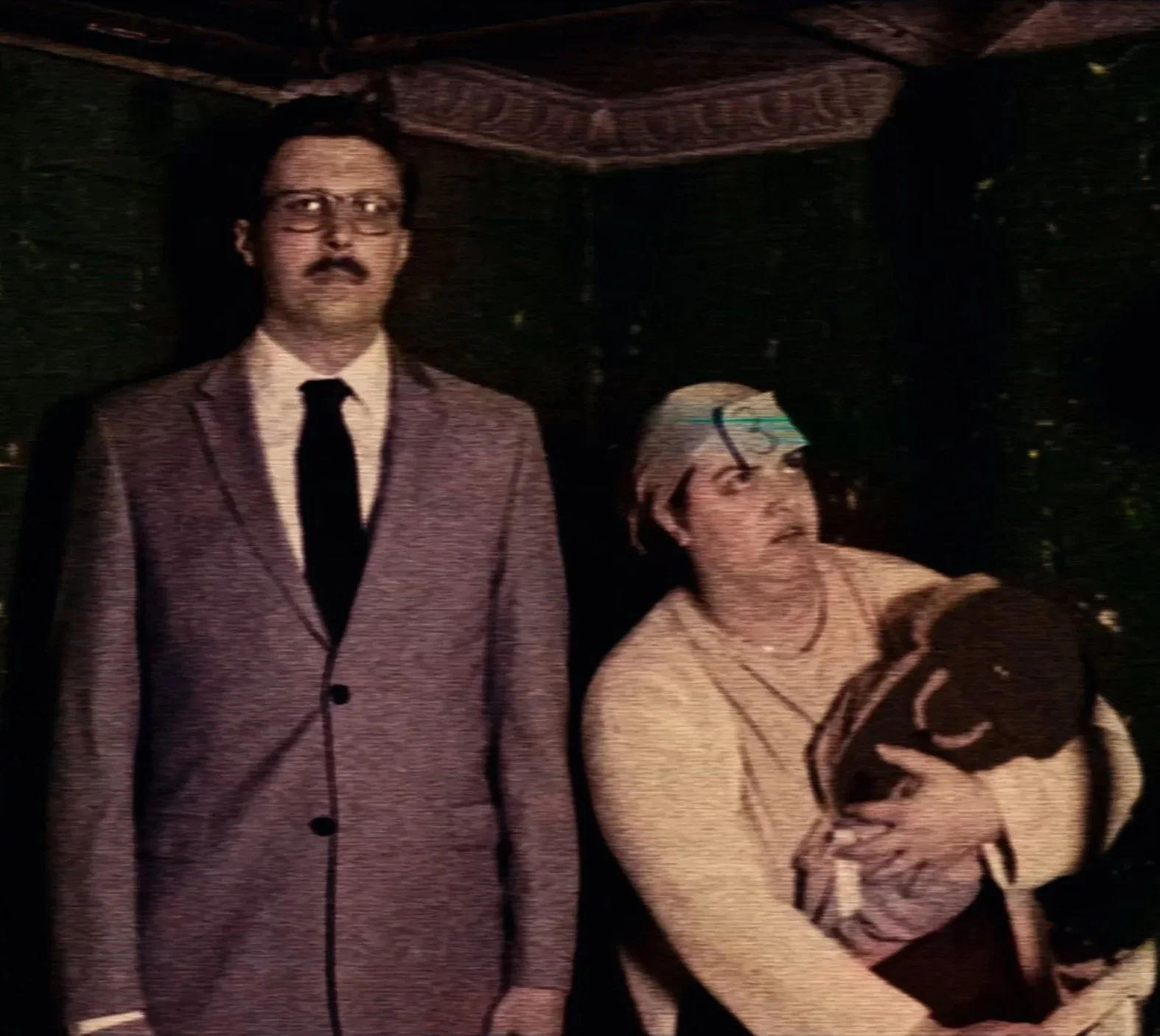 A stupid woman holding a baby in a flap eared cap and a man in a suit with glasses, stand in a dingy basement. Drew Gregory & Lexi Costantini