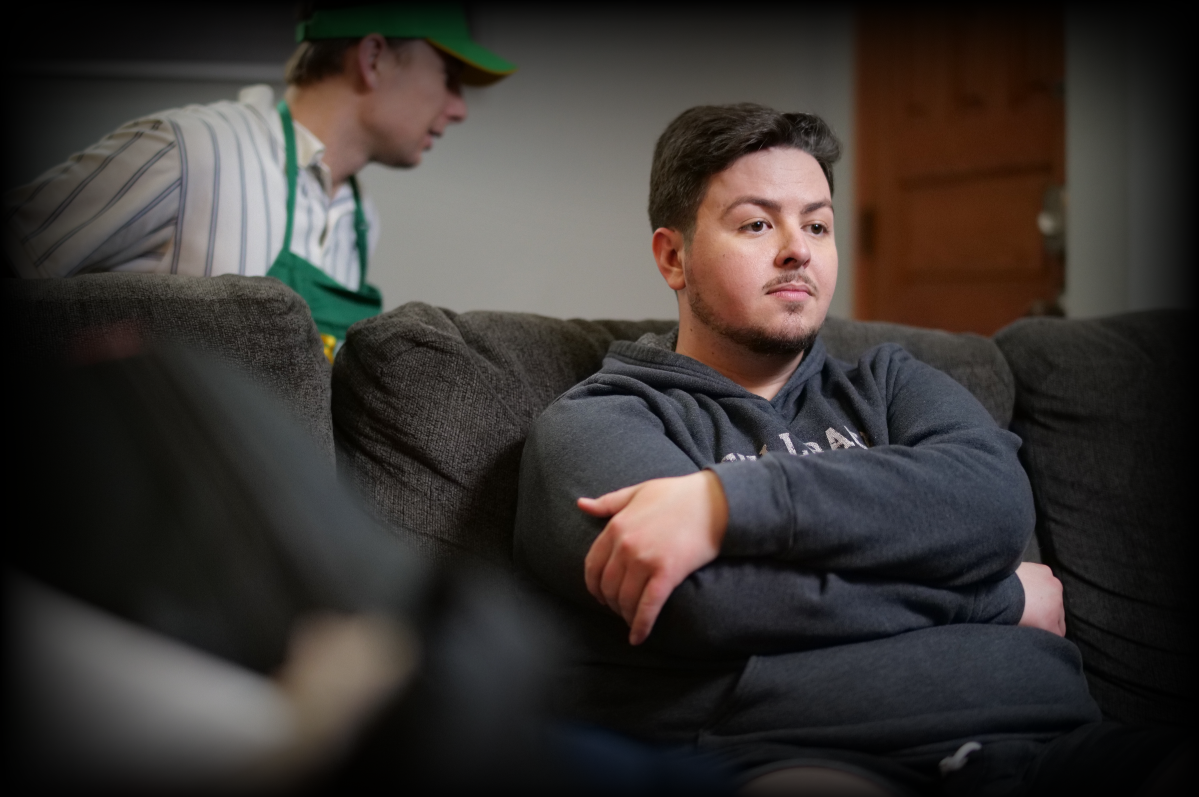 A young man with dark hair and a beard, wearing a gray hoodie, sitting on a dark gray couch with his arms crossed and looking to the side with a neutral expression. In the background, a person wearing a green cap, a green apron over a striped shirt, is leaning forward and talking to him in a casual indoor setting.